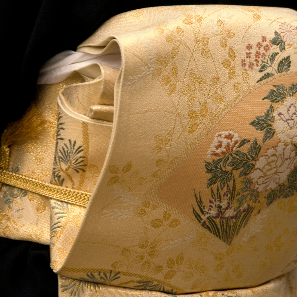 Détail d’un kimono avec obi vintage en soie jaune pâle, orné de broderies florales délicates représentant des chrysanthèmes et feuillages, avec des reflets dorés et une texture raffinée.