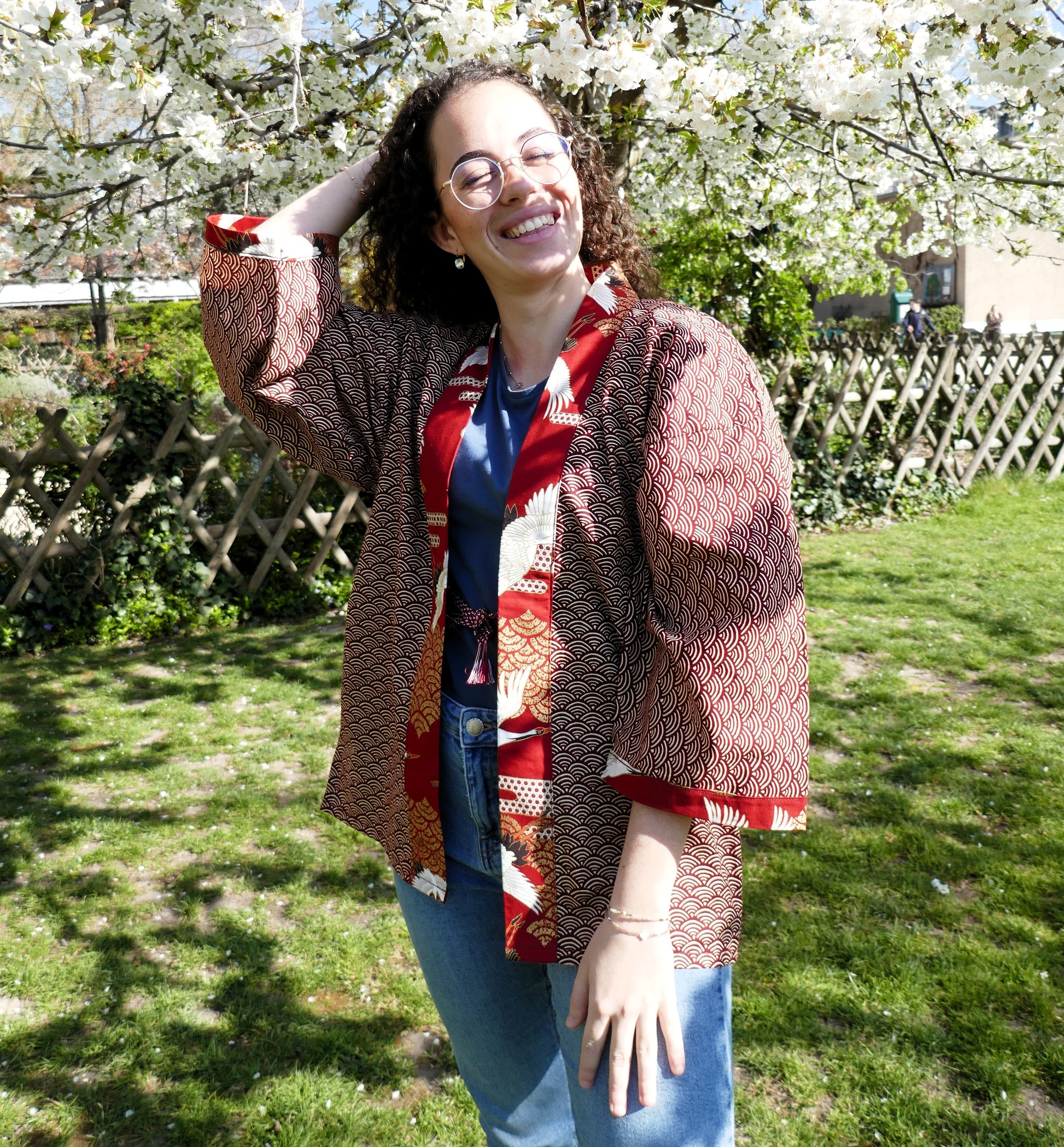Loony porte le haori japonais Akane Tsuru, motif seigaiha rouge et grues, devant des sakura pendant le hanami. Look doux et moderne.