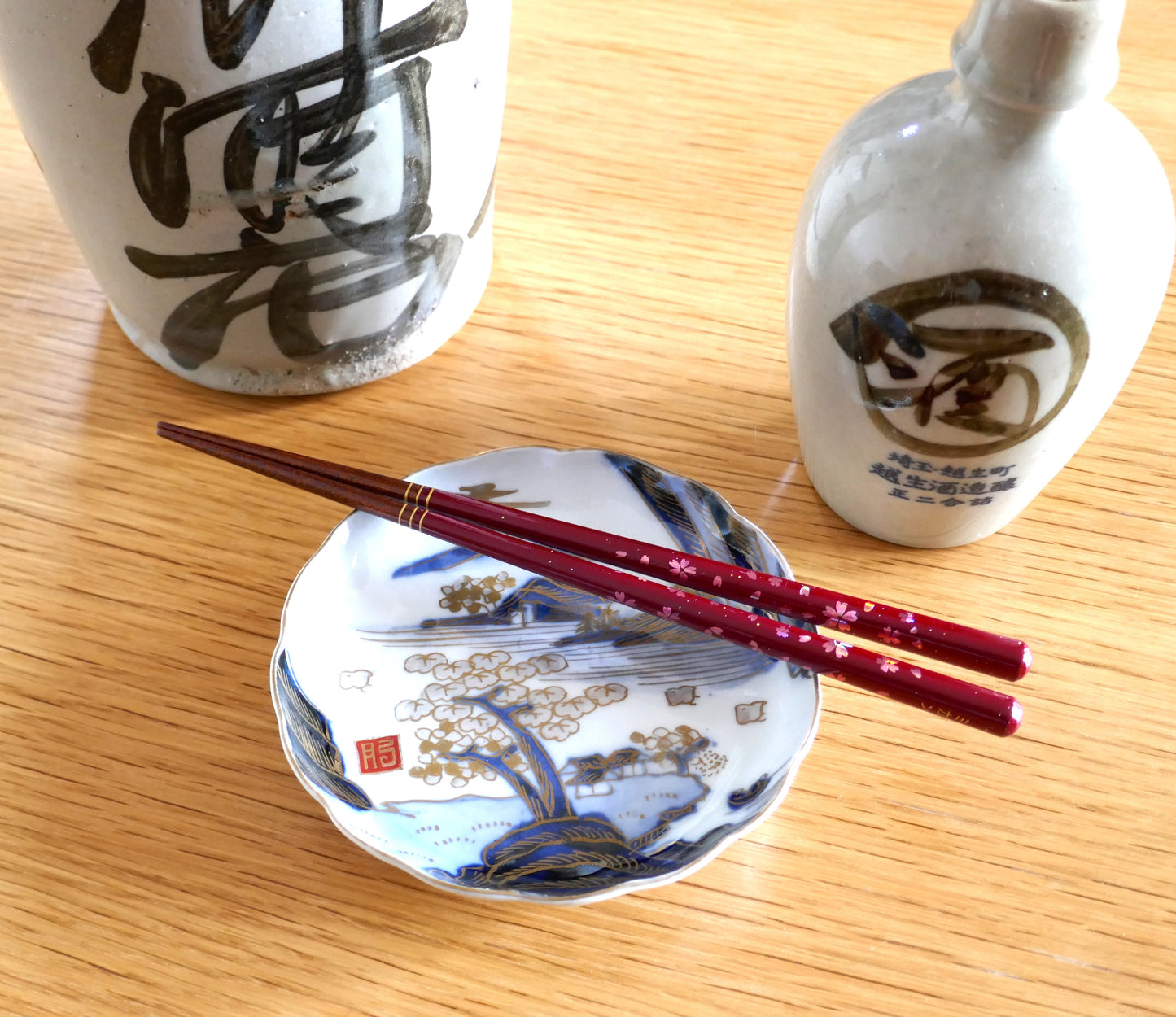 Mise en scène d’une assiette Arita Meiji avec baguettes japonaises sur fond bois et flacons de saké