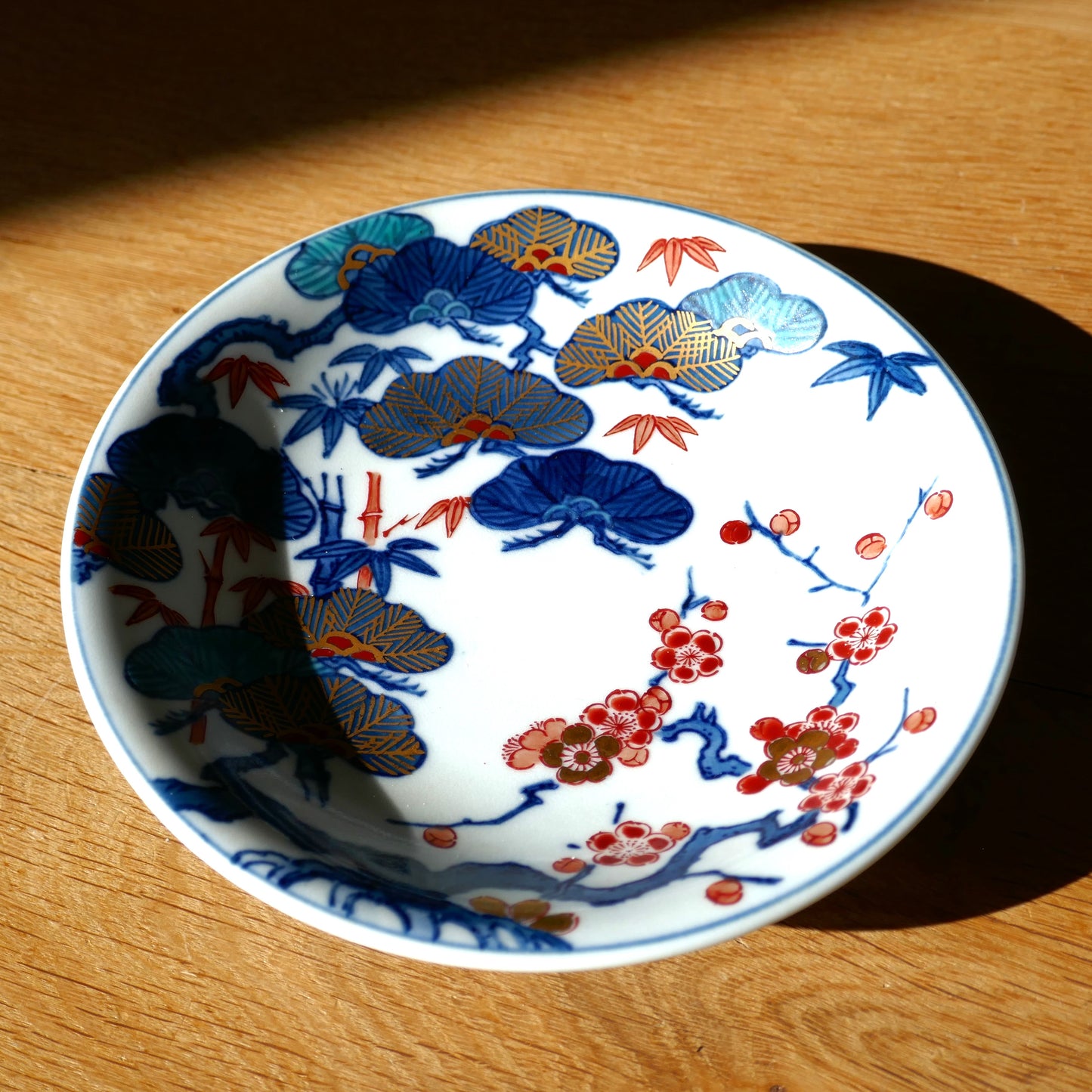 Assiette japonaise ancienne en porcelaine Arita Imari, vue partielle en lumière rasante sur table en bois. Décor peint à la main de fleurs de prunier, branches de pin et feuilles de bambou en rouge, bleu et or.