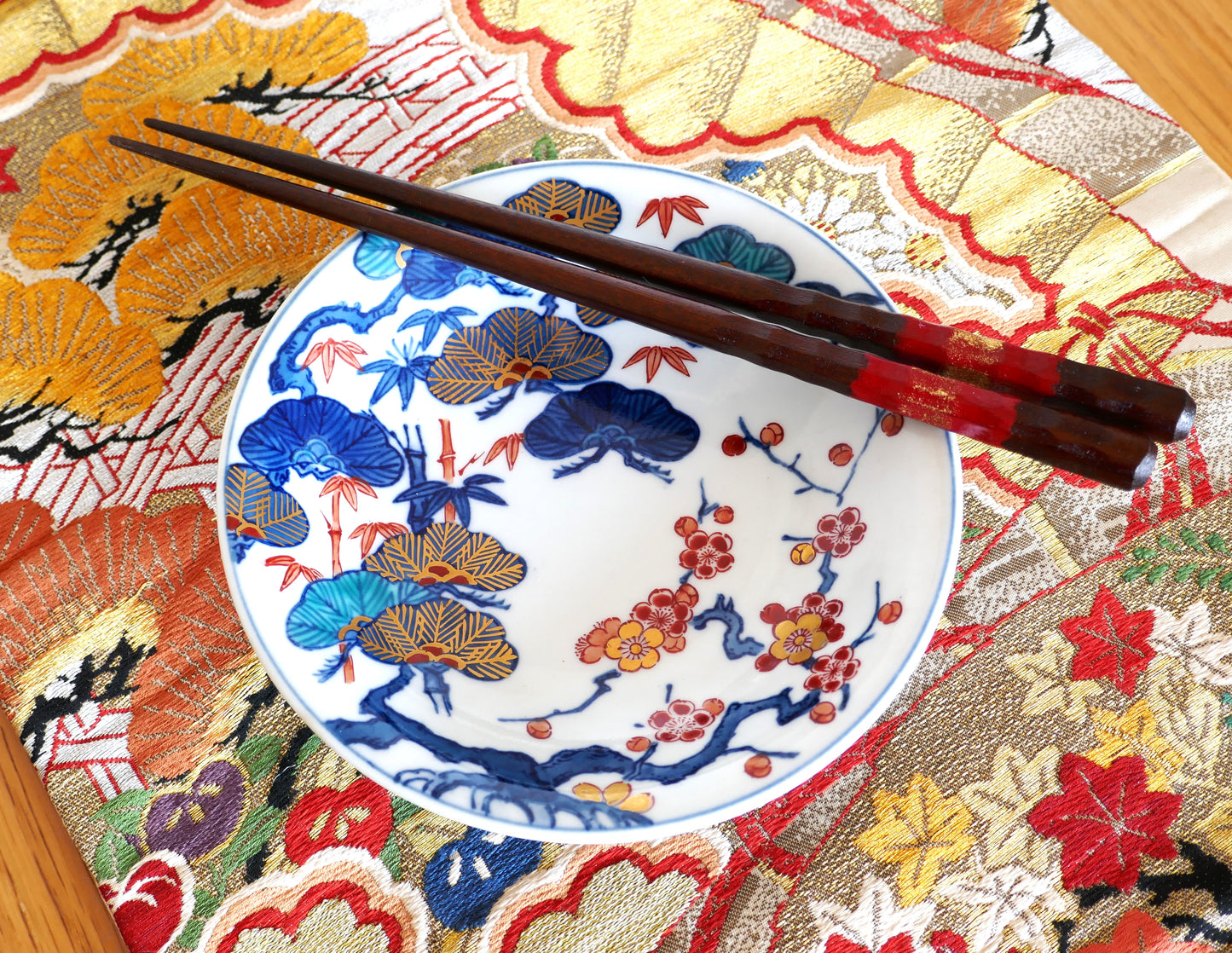 Assiette en porcelaine japonaise Imari de style Arita, décor polychrome traditionnel Sho Chiku Bai (pin, bambou et prunier). Présentation sur un tissu obi brodé aux motifs floraux dorés, avec baguettes en bois laqué rouge et or.
