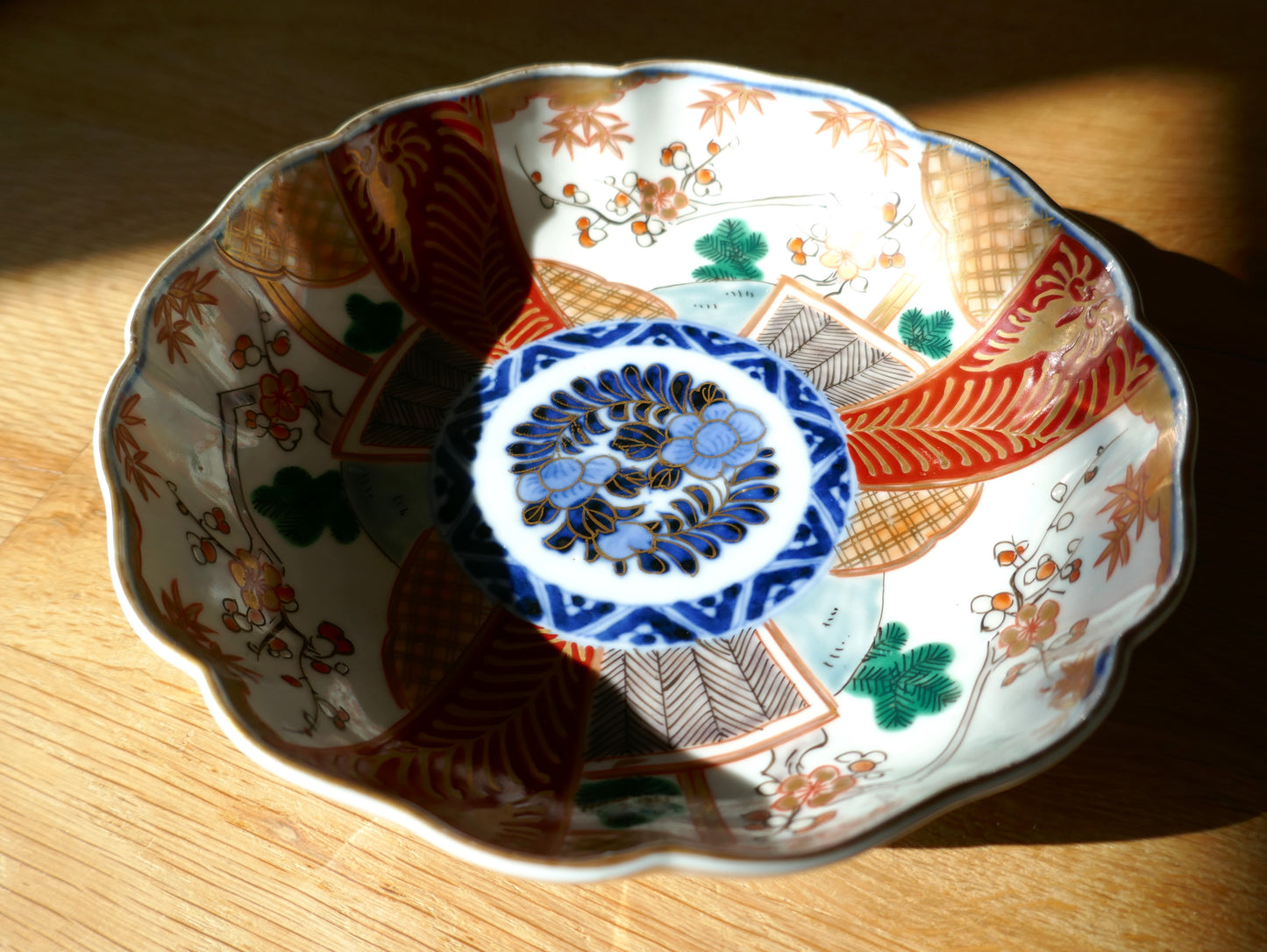 Grande assiette ancienne en porcelaine japonaise Imari Meiji, à décor segmenté rouge, vert, or et bleu cobalt, illuminée par une lumière rasante. Médaillon central floral cerné de motifs géométriques et floraux traditionnels.