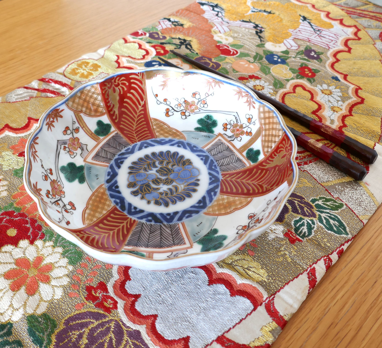 Grande assiette japonaise en porcelaine Imari de la période Meiji, au décor polychrome segmenté avec médaillon central floral bleu, rouge et or. Présentée avec des baguettes laquées sur un tissu brocart obi aux motifs traditionnels japonais.
