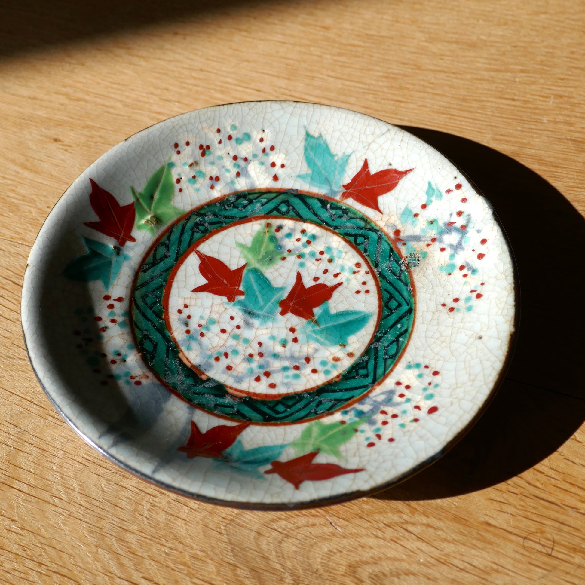Assiette ancienne japonaise en céramique Inuyama-yaki, décorée à la main de momiji (feuilles d’érable) rouges et turquoise. Céramique vernissée à glaçure craquelée typique, mise en lumière naturelle sur fond en bois.