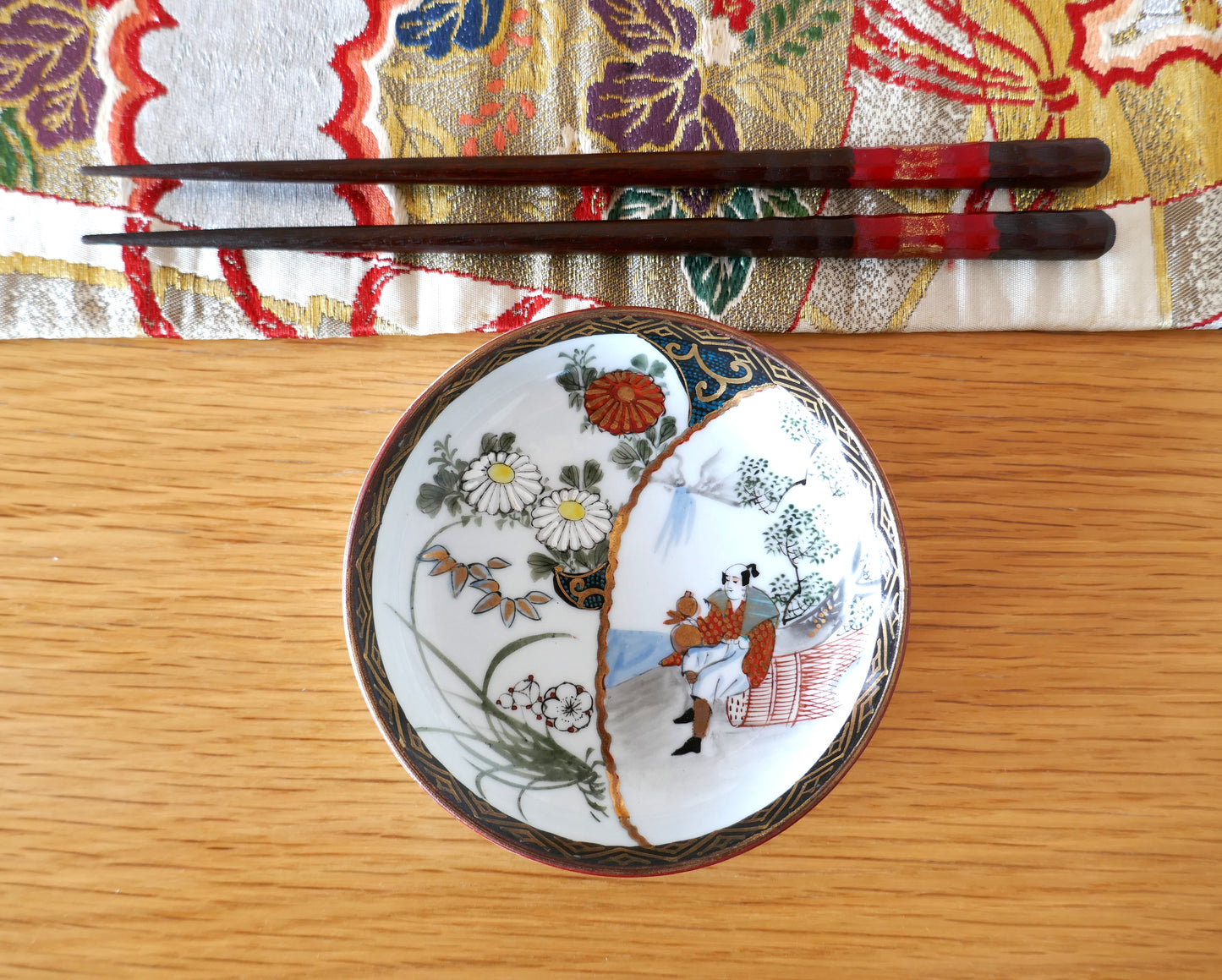 Assiette Kutani avec décor floral et scène figurative, accompagnée de baguettes japonaises sur une table en bois