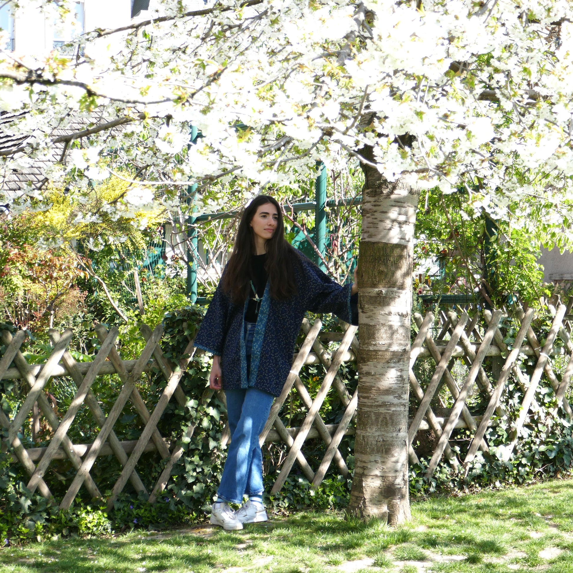 Mayssa porte le haori Full Tombo sous un cerisier en fleurs, silhouette élégante et tissu bleu marine à motifs de libellules.