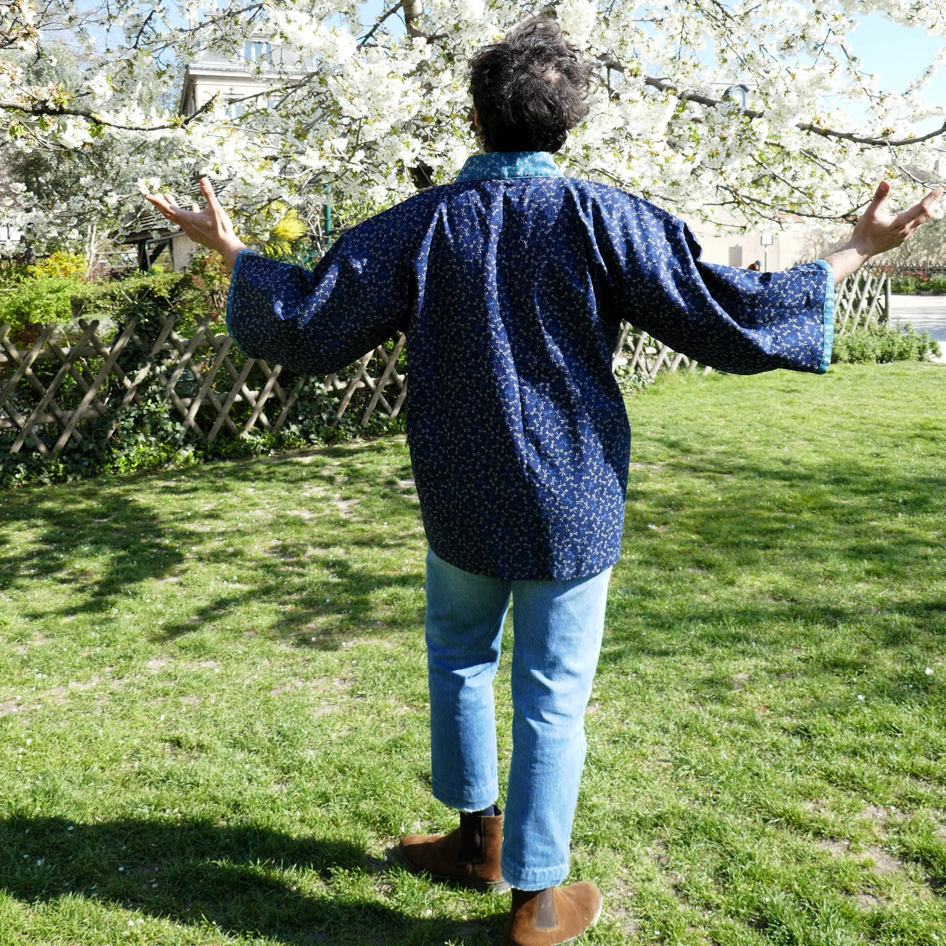 Richard porte le haori japonais Full Tombo de l'atelier KOGEDO vu de dos, bras ouverts sous un cerisier en fleurs, tissu bleu à motif de libellules.