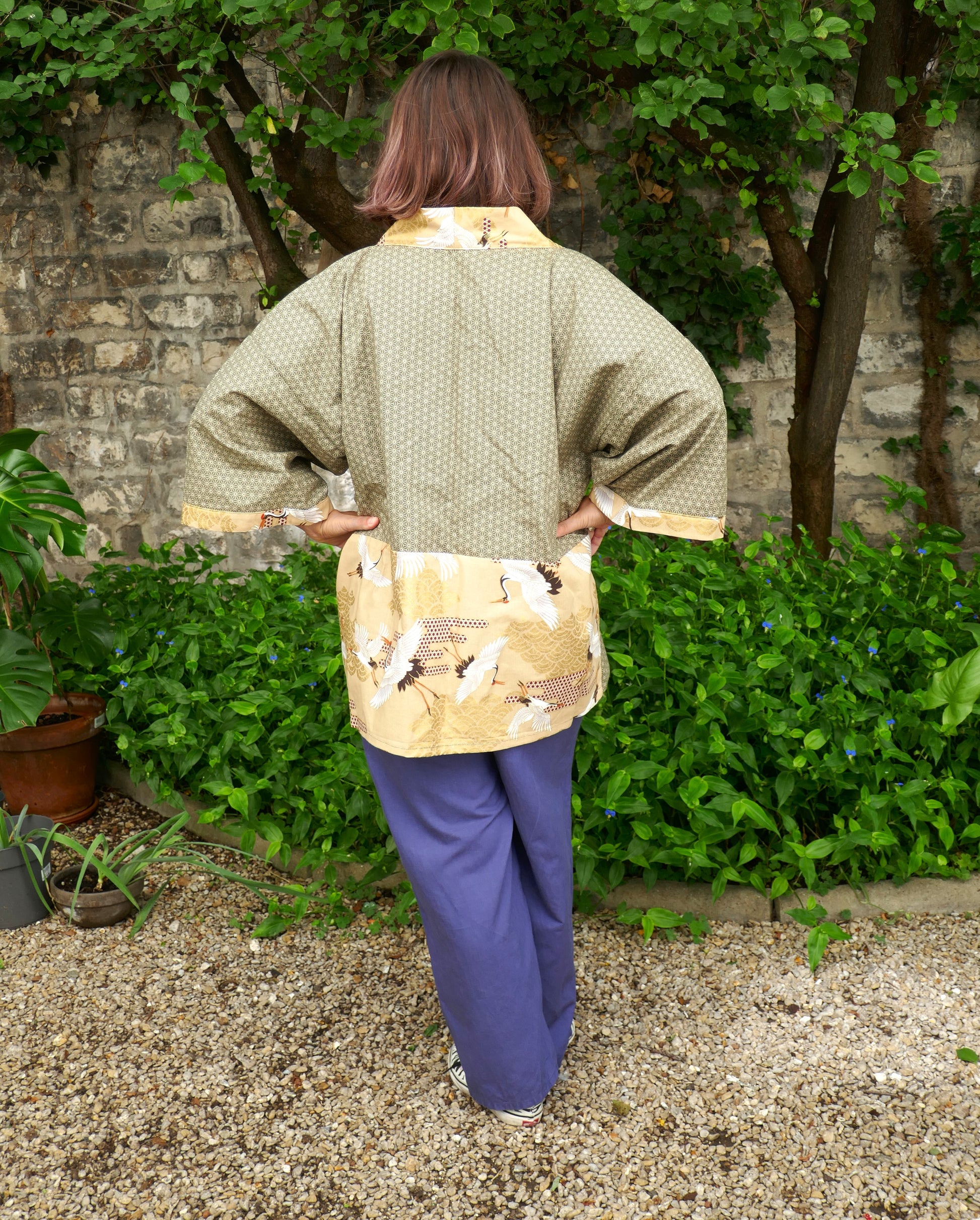 Mikan vue de dos portant un haori kimono de l’atelier KOGEDO, modèle "Nohara no Tsuru". Veste verte à motif asanoha et empiècements beiges imprimés de grues japonaises, ambiance naturelle et mur en pierre en arrière-plan.