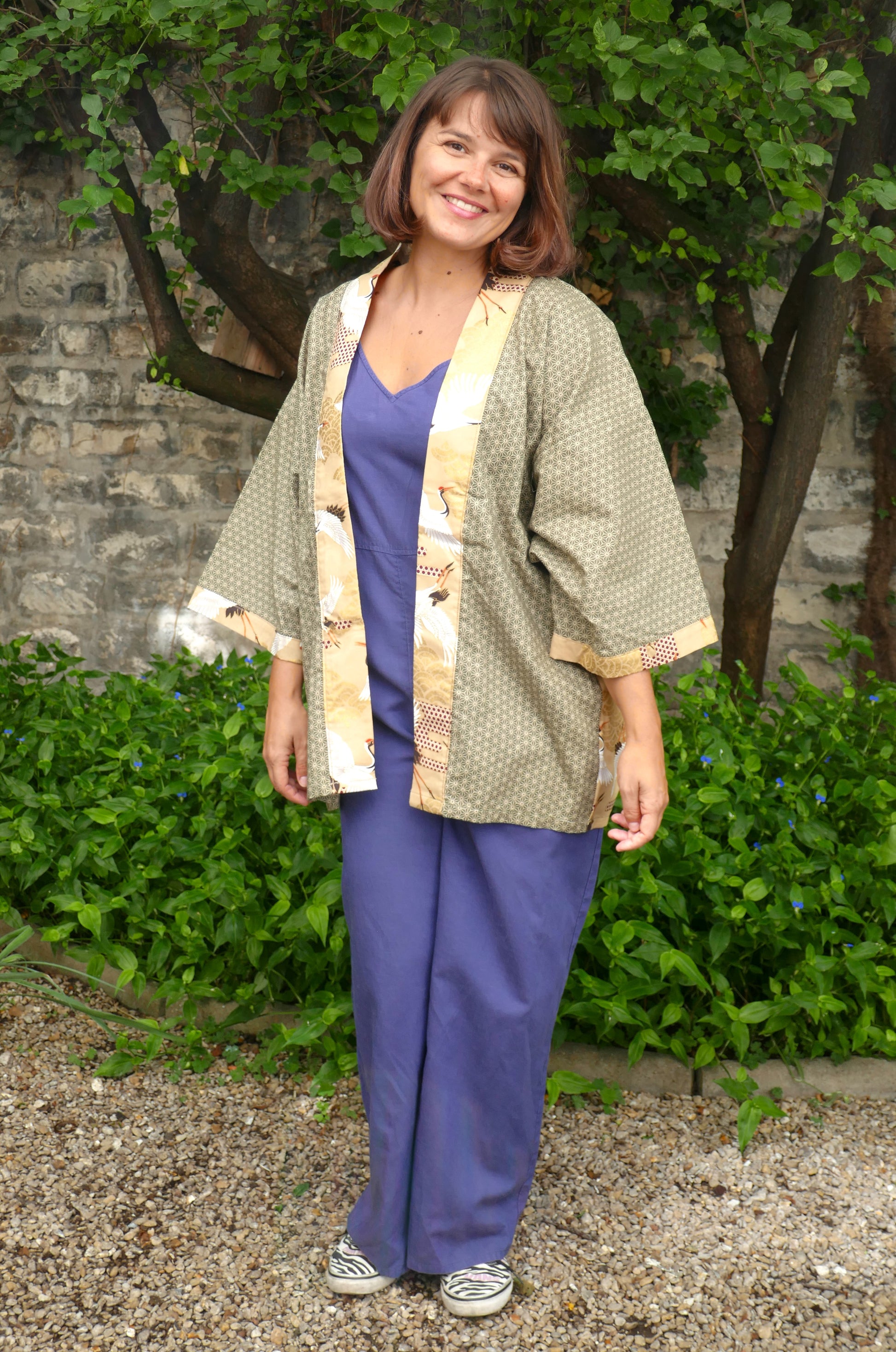 Mikan souriante vêtue du haori unisexe "Nohara no Tsuru" signé KOGEDO, avec col et bords beiges imprimés de grues et fond vert motif géométrique japonais. Style bohème japonais moderne dans un jardin fleuri.