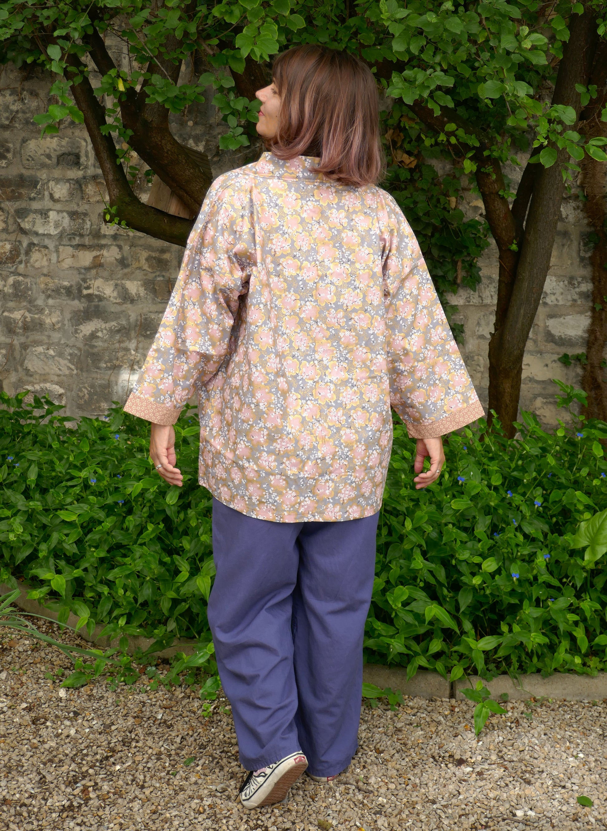 Mikan vue de dos dans le haori moderne “Sakura Fubuki” de l’atelier KOGEDO. Veste kimono japonaise légère au motif floral rose et pêche sur fond gris, ambiance poétique et printanière en extérieur.