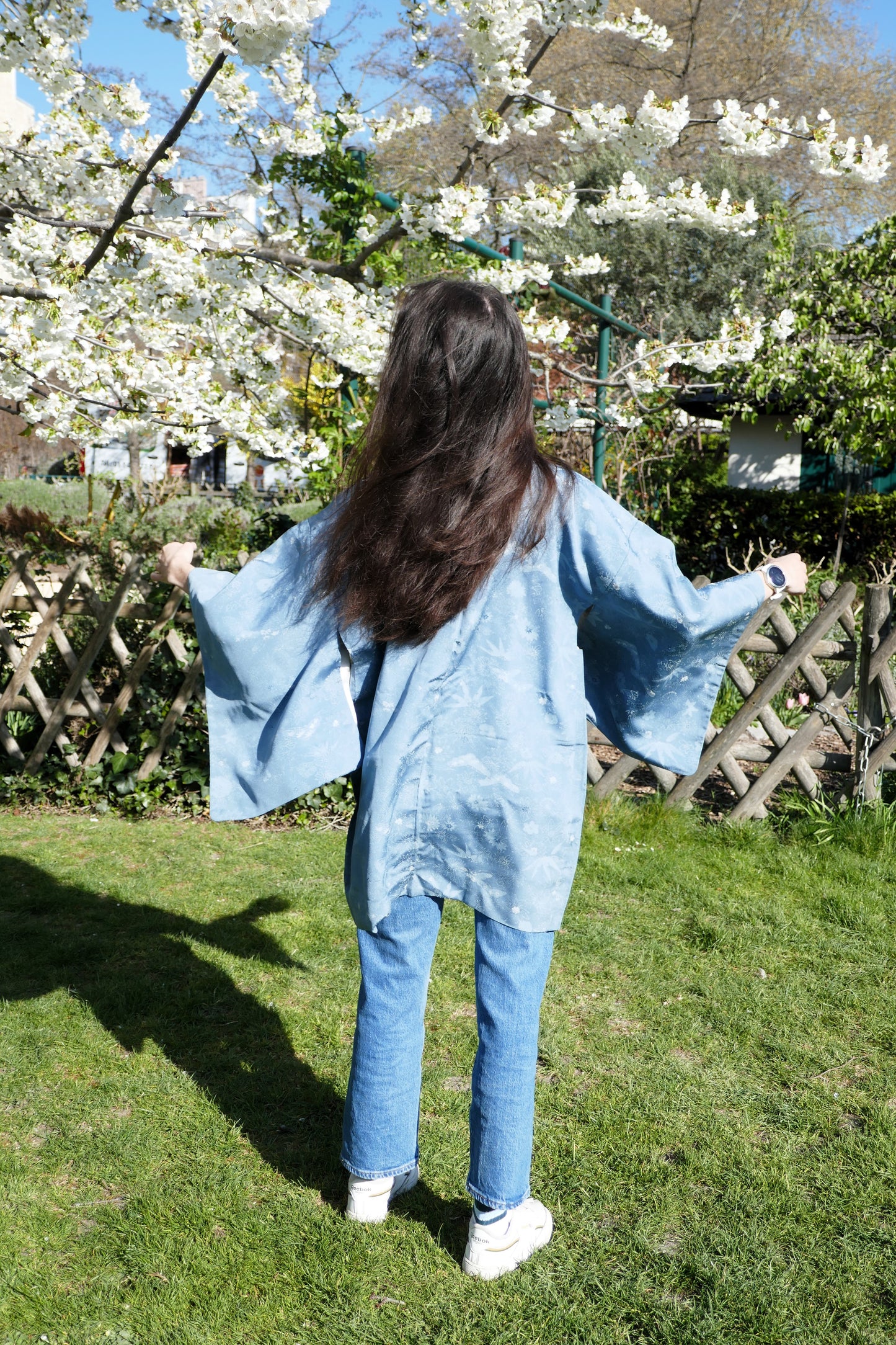 Vue de dos du haori bleu en soie porté par Mayssa, manches déployées devant des fleurs de cerisier
