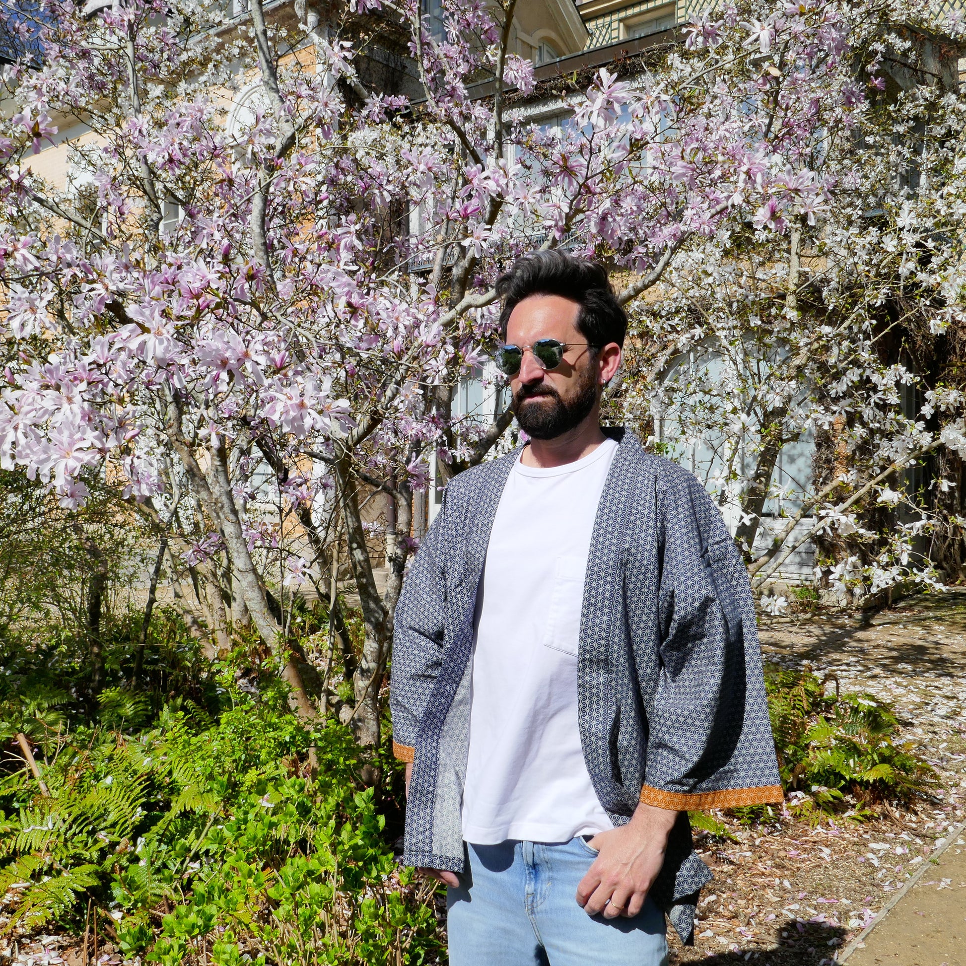 Tôma porte le haori Tradi Tradi de KOGEDO dans un jardin en fleurs, coupe oversize tombante sur un tee-shirt blanc et un jean clair.