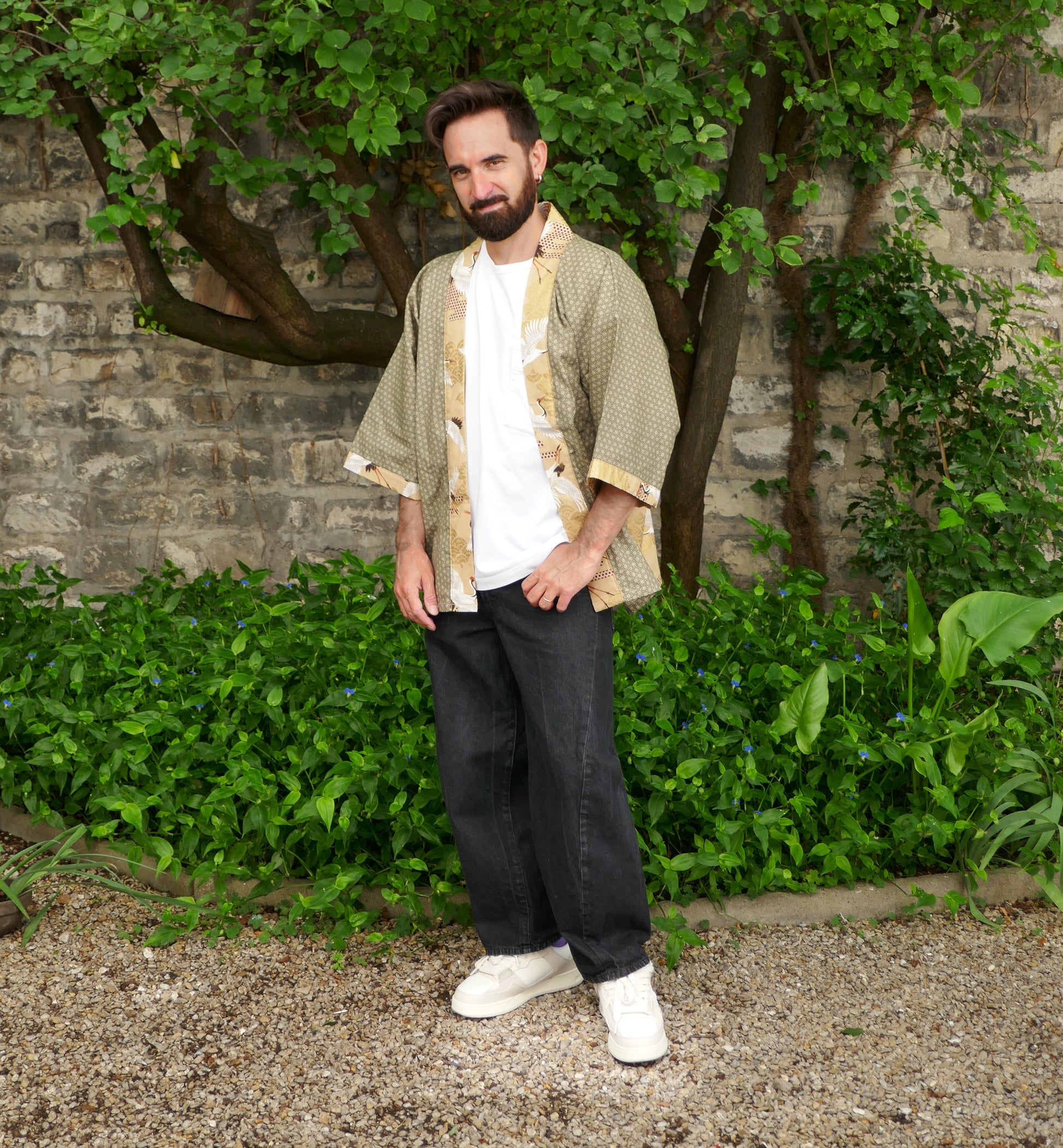 Toma portant un haori japonais moderne, création KOGEDO, modèle "Nohara no Tsuru" vert kaki et beige à motifs de grues et d’asanoha. Look décontracté avec t-shirt blanc, jean noir et baskets blanches, dans un jardin naturel.