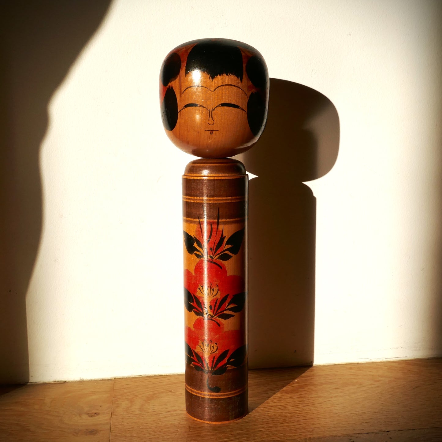 Kokeshi japonaise Shingata artisanale par Kiyohara Takao, décor de camélias rouges et feuilles noires, vue sous lumière naturelle avec ombres