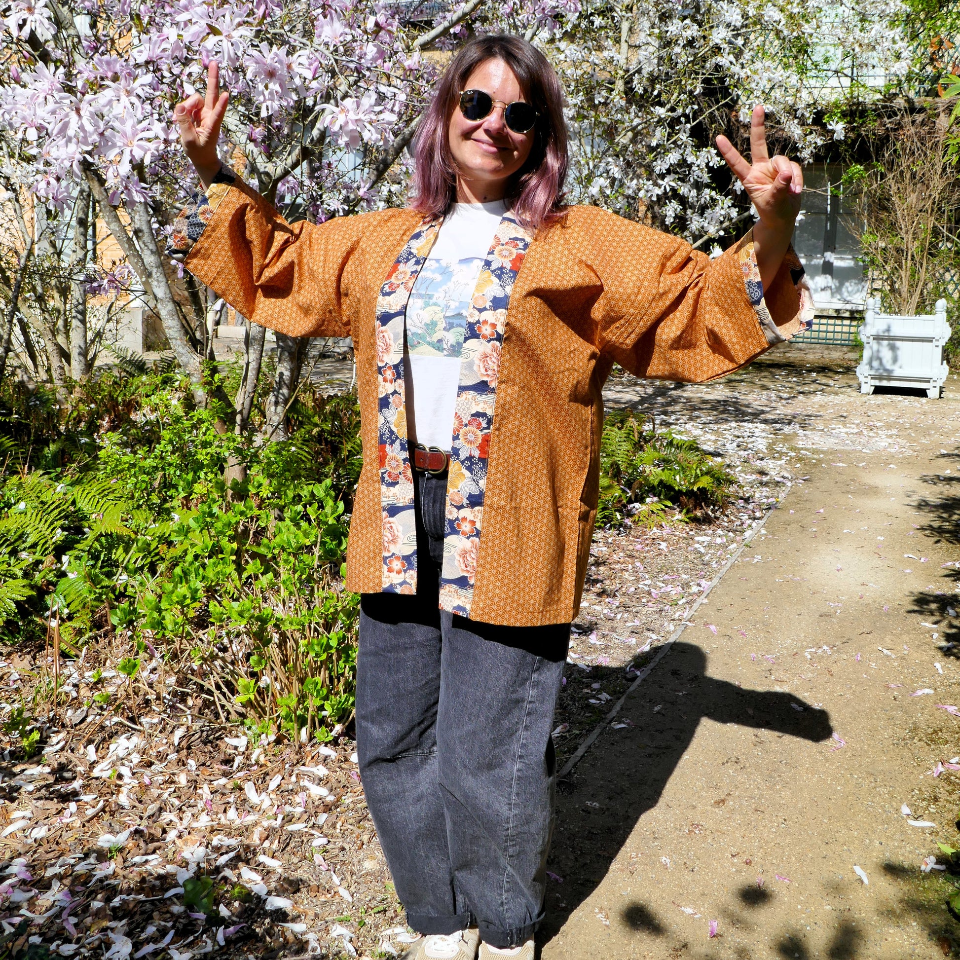 Mikan souriante en haori Yūyake Zakura, sous un magnolia en fleurs, faisant le signe de paix.