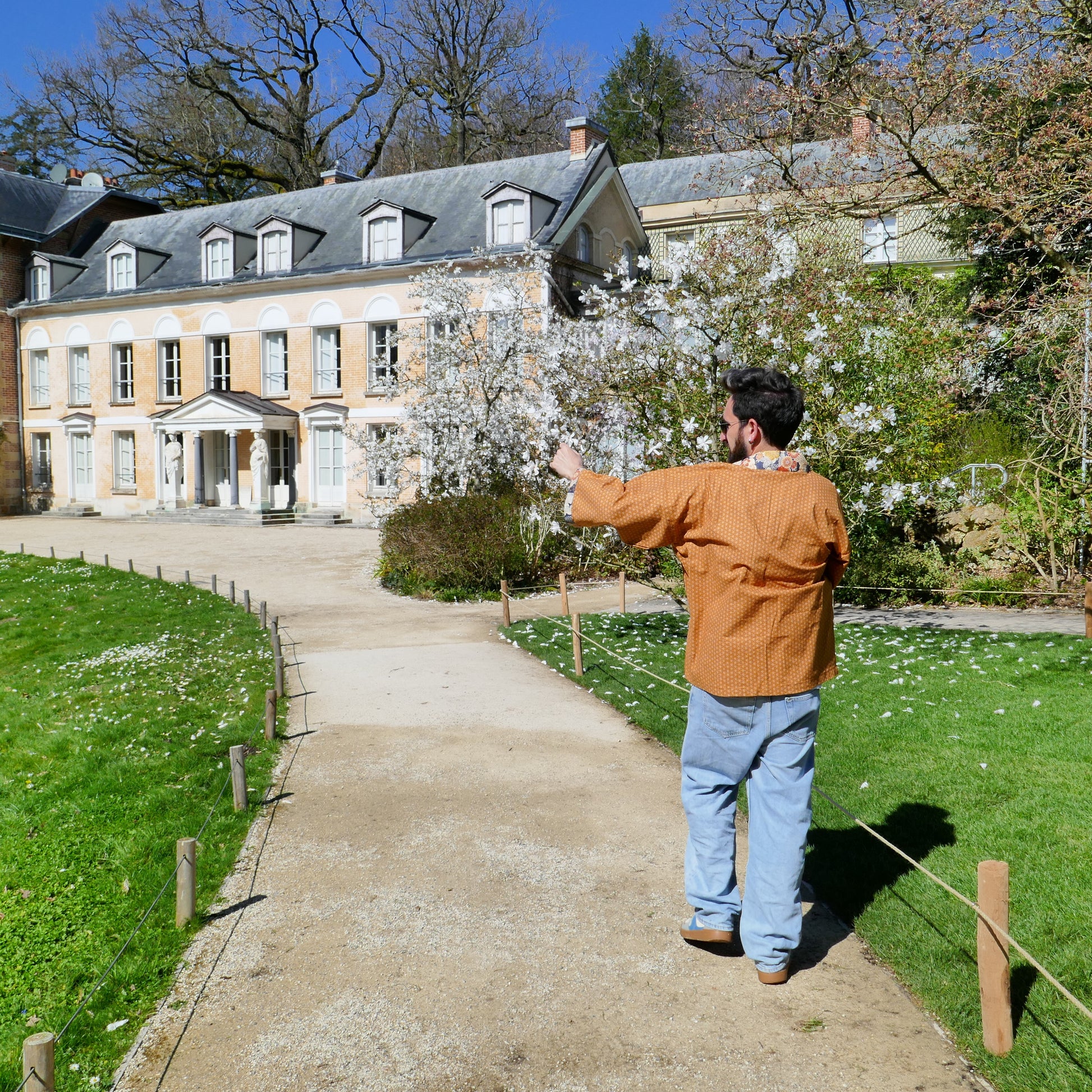 Tôma vu de dos en haori Yūyake Zakura, marchant vers un grand cerisier devant une maison ancienne.