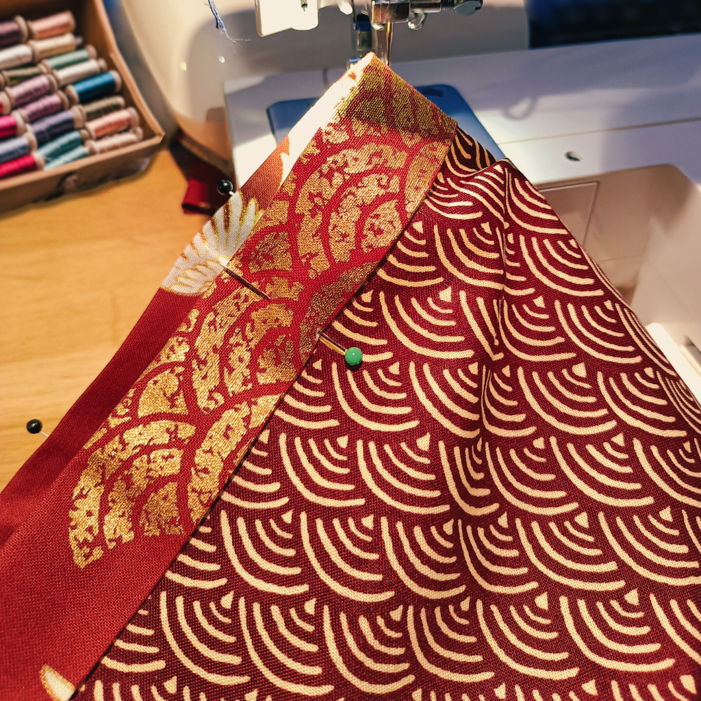 Tissu japonais rouge orné de motifs Seigaiha dorés en cours de confection sur une machine à coudre, dans un atelier artisanal. En arrière-plan, des bobines de fil multicolores illustrent le travail minutieux et créatif du haori fait main.