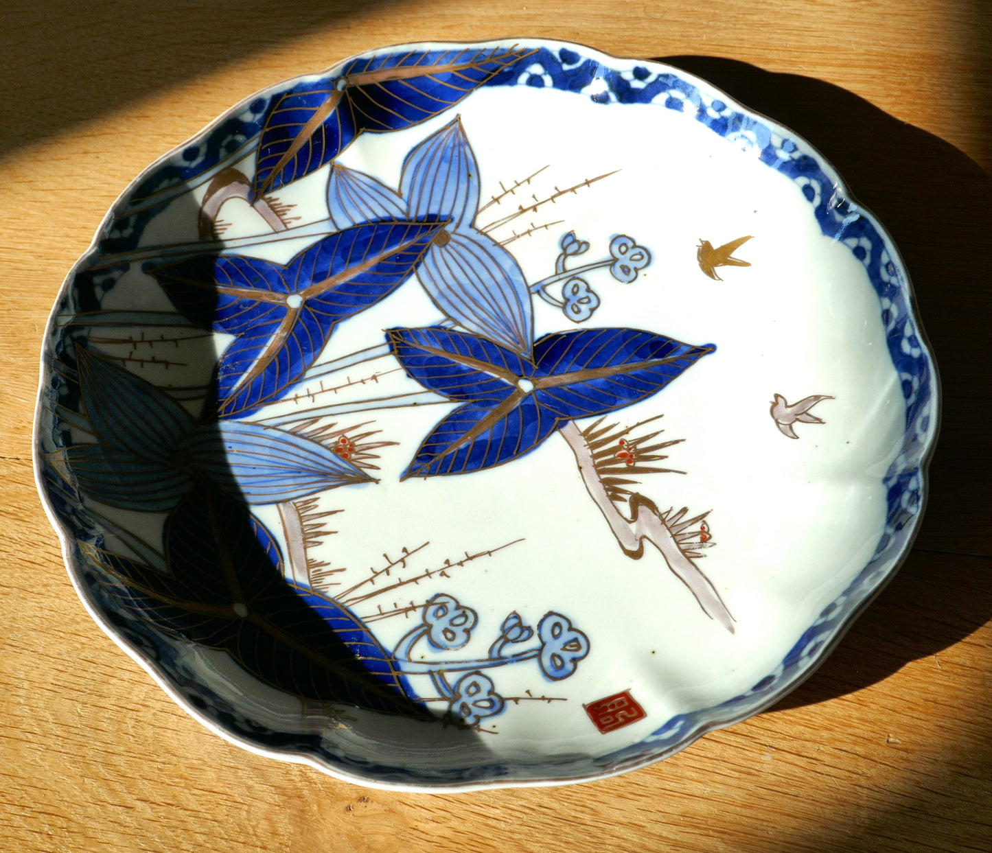 Assiette japonaise en porcelaine d’Arita, époque Meiji, vue partielle à la lumière rasante. Décor floral de feuilles bleu indigo et or, style traditionnel japonais sur faïence blanche à bord festonné.