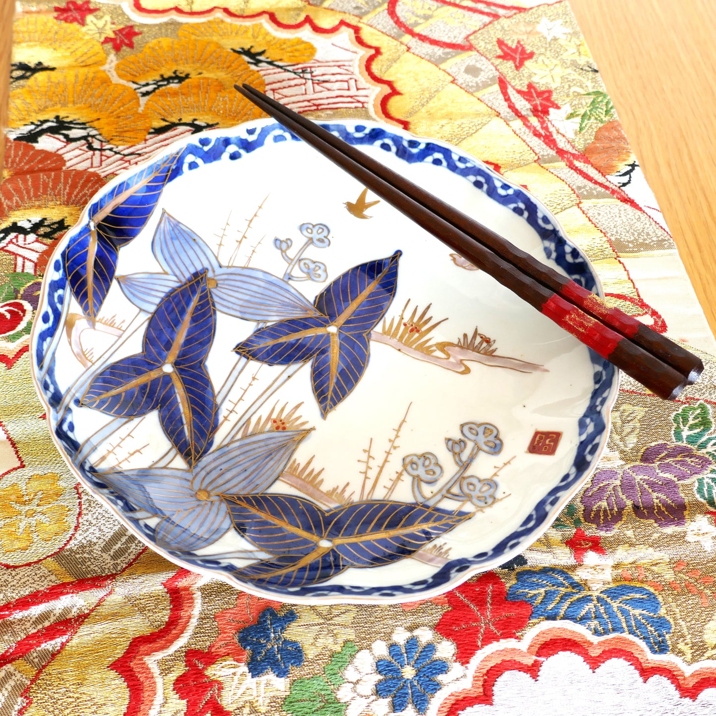 Assiette en porcelaine japonaise Arita Meiji posée sur un obi en soie brodée, accompagnée de baguettes japonaises en bois laqué rouge. Décor raffiné de feuilles et oiseaux dorés, esthétique traditionnelle.