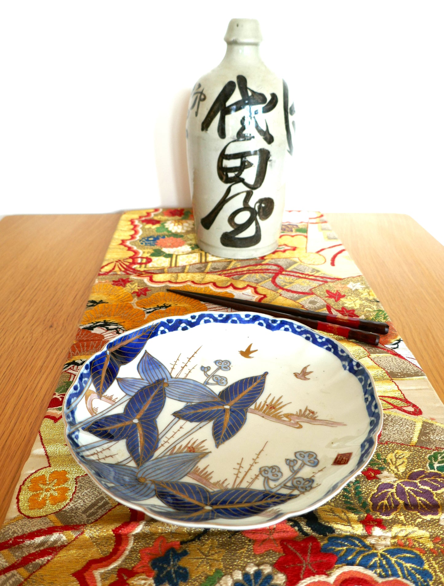 Mise en scène d’une assiette Arita Meiji en porcelaine décorée de feuilles bleues et d’oiseaux, accompagnée d’une bouteille de saké calligraphiée. Présentation sur un obi brodé, ambiance authentique et élégante.