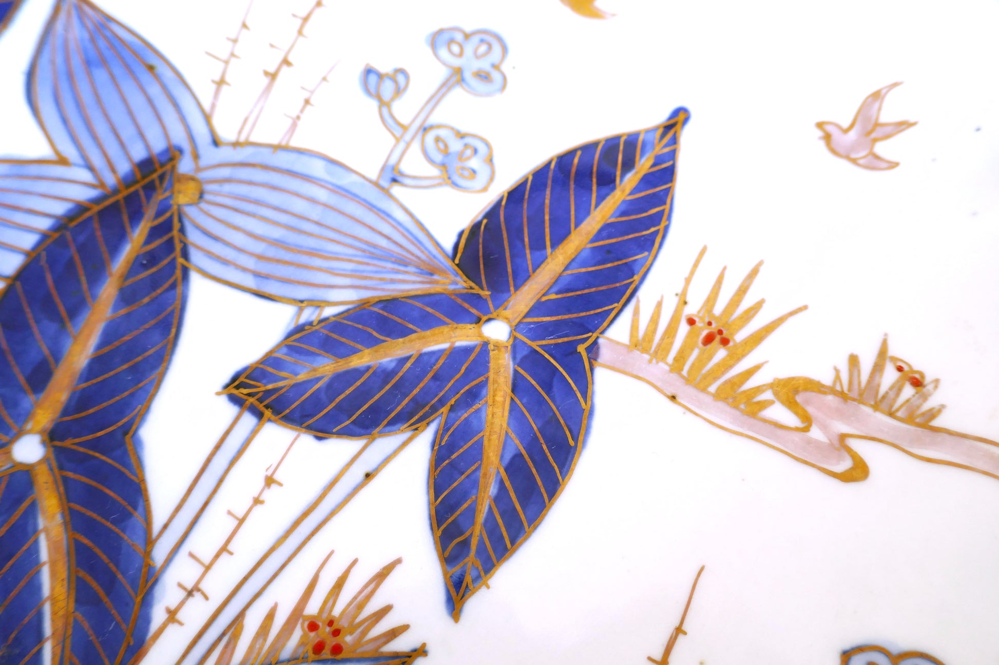 Gros plan sur une assiette en porcelaine japonaise Arita de la période Meiji, décorée de feuilles bleues stylisées et de rehauts dorés sur fond blanc. Détails fins de peinture à la main représentant un motif végétal traditionnel japonais.