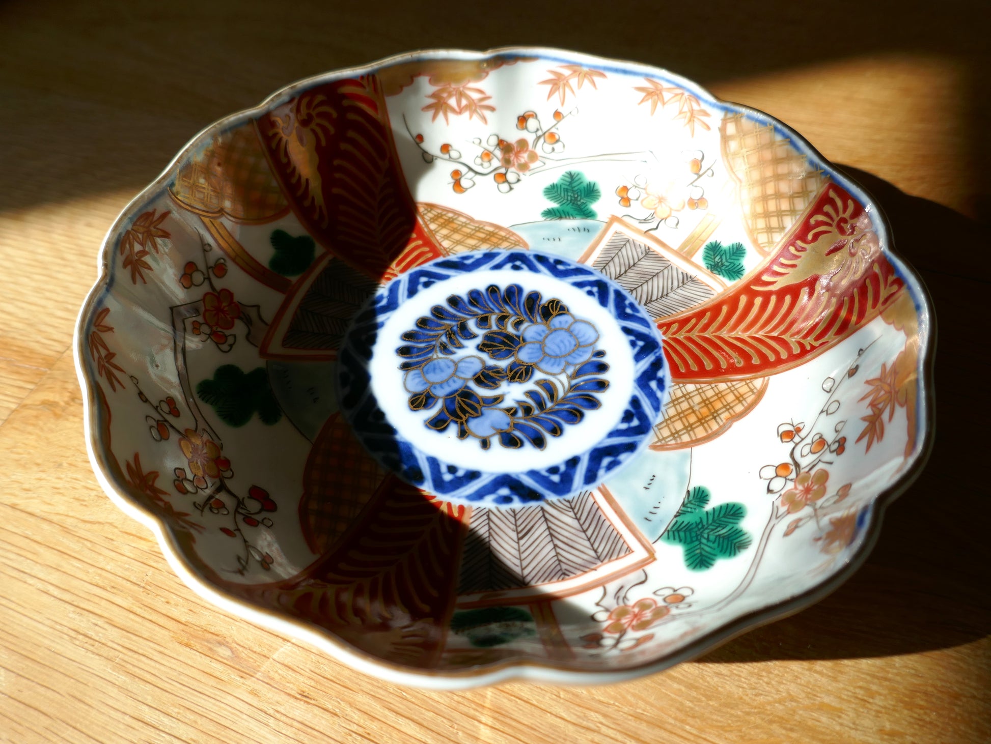 Grande assiette ancienne en porcelaine japonaise Imari Meiji, à décor segmenté rouge, vert, or et bleu cobalt, illuminée par une lumière rasante. Médaillon central floral cerné de motifs géométriques et floraux traditionnels.