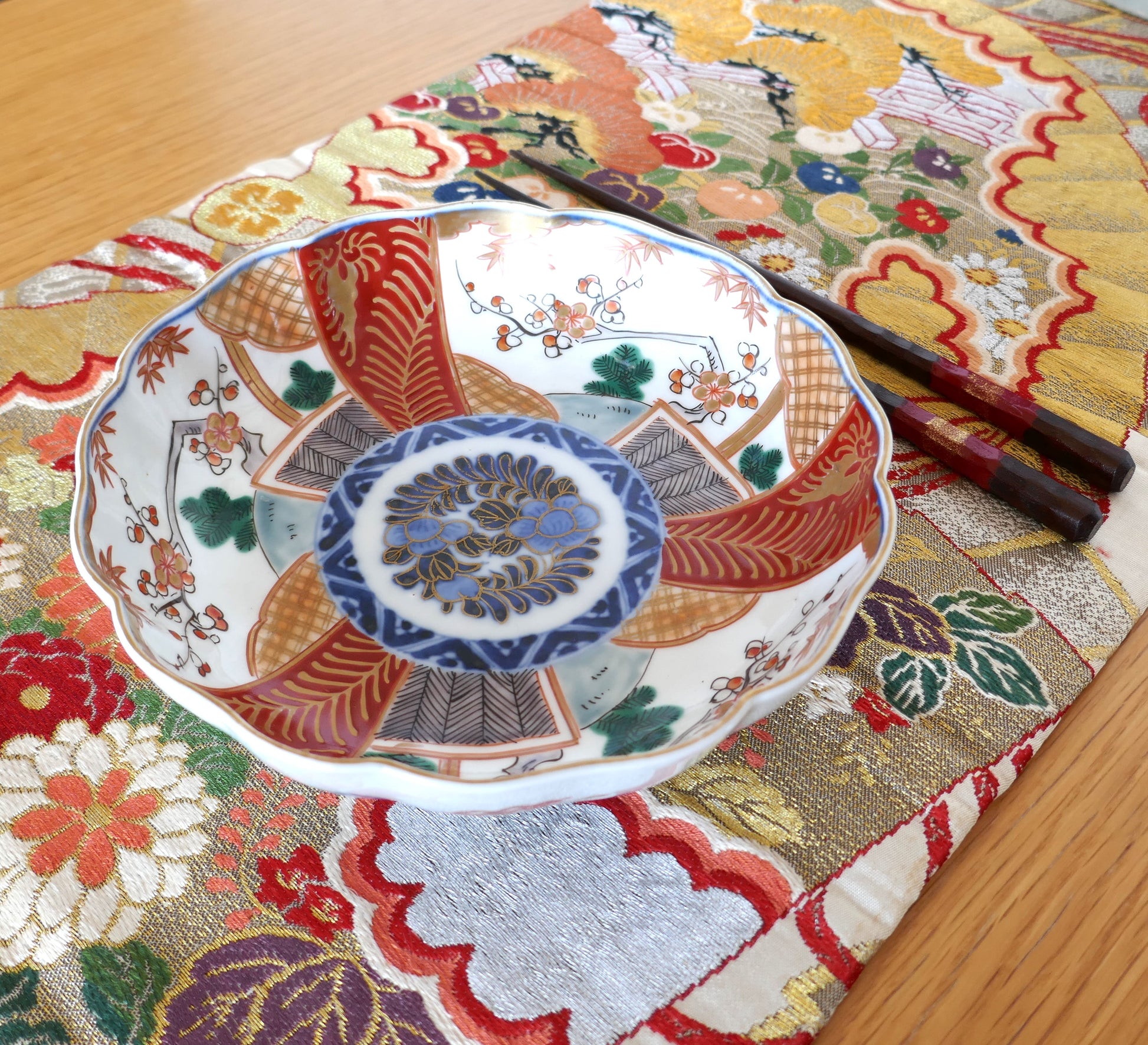 Grande assiette japonaise en porcelaine Imari de la période Meiji, au décor polychrome segmenté avec médaillon central floral bleu, rouge et or. Présentée avec des baguettes laquées sur un tissu brocart obi aux motifs traditionnels japonais.
