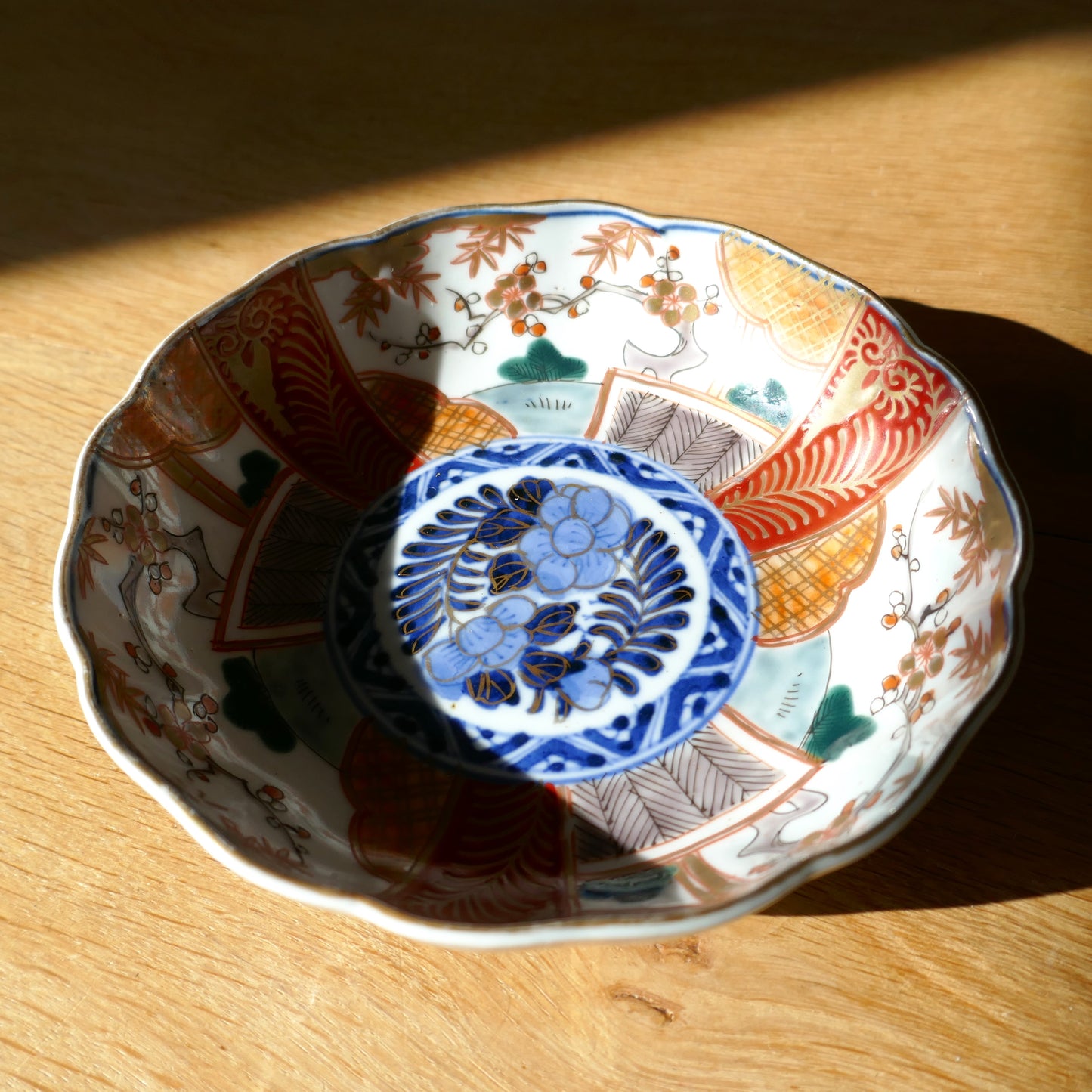 Assiette ancienne Imari de l’ère Meiji, porcelaine fine avec décor segmenté en rouge, or et vert, médaillon central bleu aux fleurs stylisées. Céramique japonaise éclairée par une lumière rasante, posée sur table en bois clair.