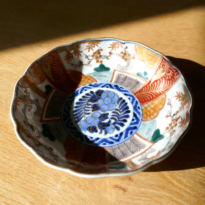 Assiette ancienne Imari de l’ère Meiji, porcelaine fine avec décor segmenté en rouge, or et vert, médaillon central bleu aux fleurs stylisées. Céramique japonaise éclairée par une lumière rasante, posée sur table en bois clair.