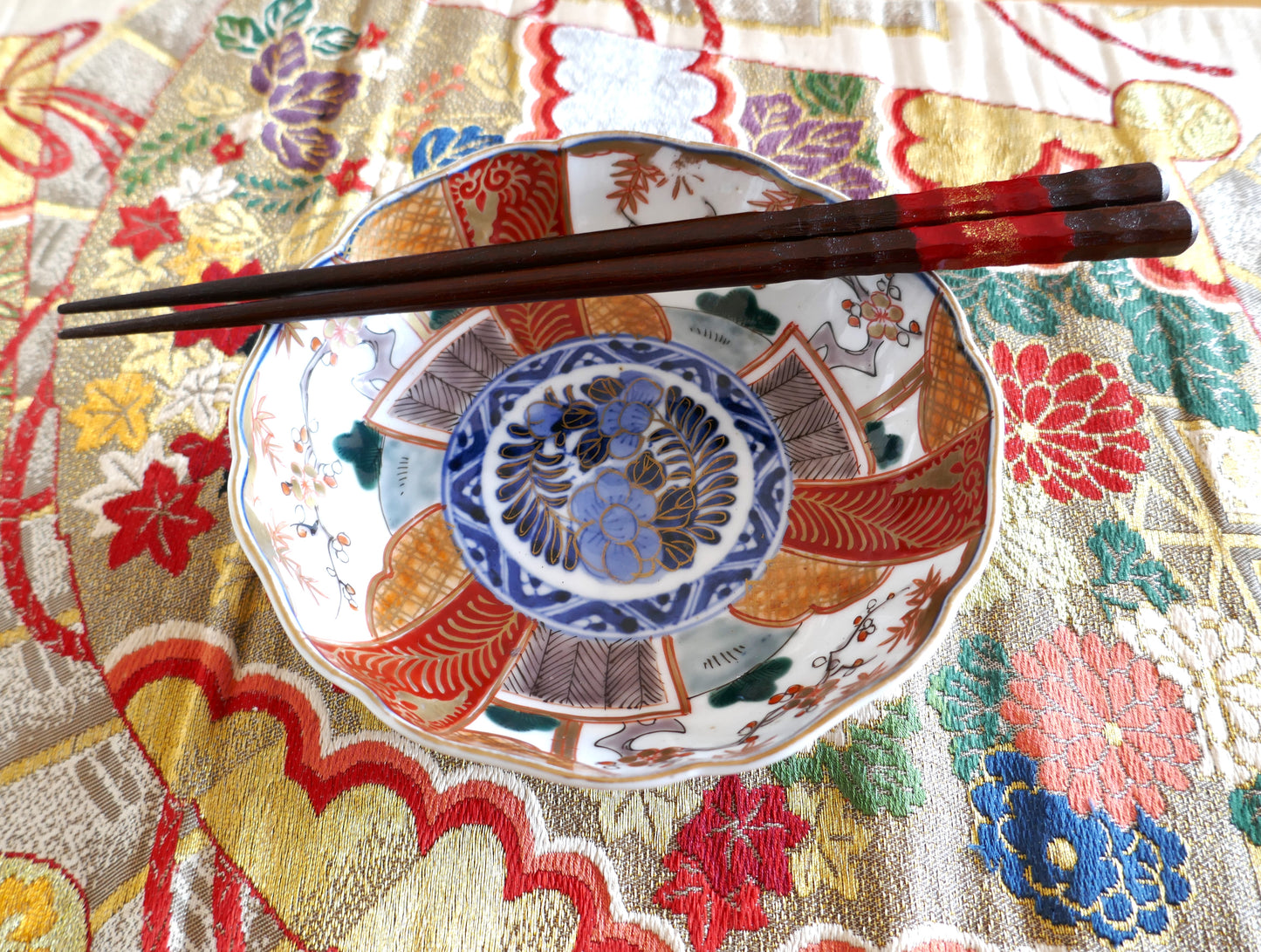 Assiette japonaise en porcelaine Imari Meiji, décorée de panneaux polychromes rouges, or et verts autour d’un médaillon central bleu cobalt. Présentée avec des baguettes laquées sur un tissu obi brodé de motifs floraux japonais.