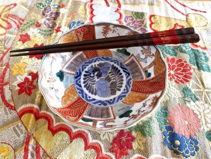 Assiette japonaise en porcelaine Imari Meiji, décorée de panneaux polychromes rouges, or et verts autour d’un médaillon central bleu cobalt. Présentée avec des baguettes laquées sur un tissu obi brodé de motifs floraux japonais.