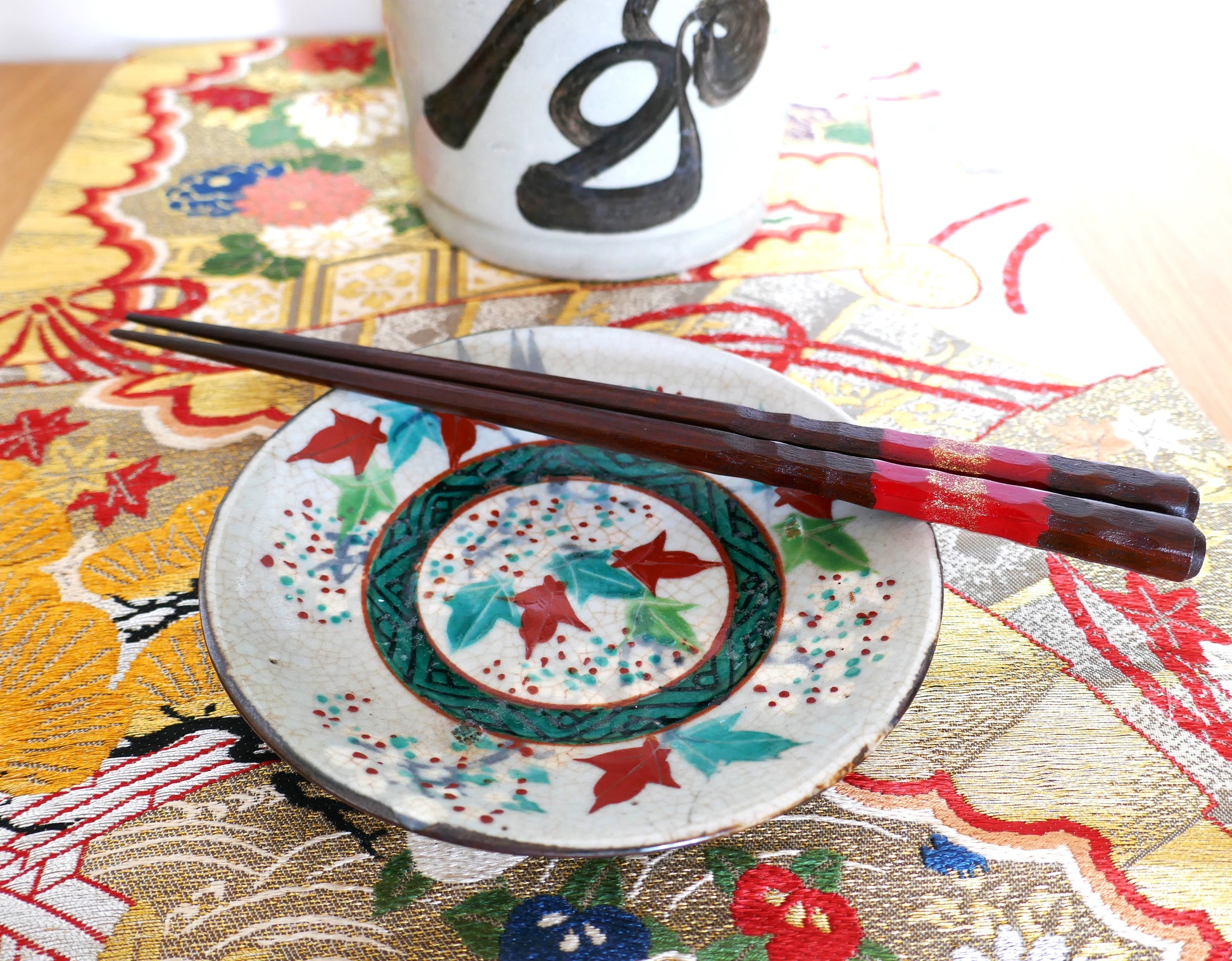 Petite assiette japonaise Inuyama-yaki de l’époque Edo, décorée de feuilles d’érable rouges et vertes sur fond craquelé. Présentée avec des baguettes en bois laqué rouge sur un obi brodé aux motifs floraux dorés et une bouteille de saké calligraphiée.