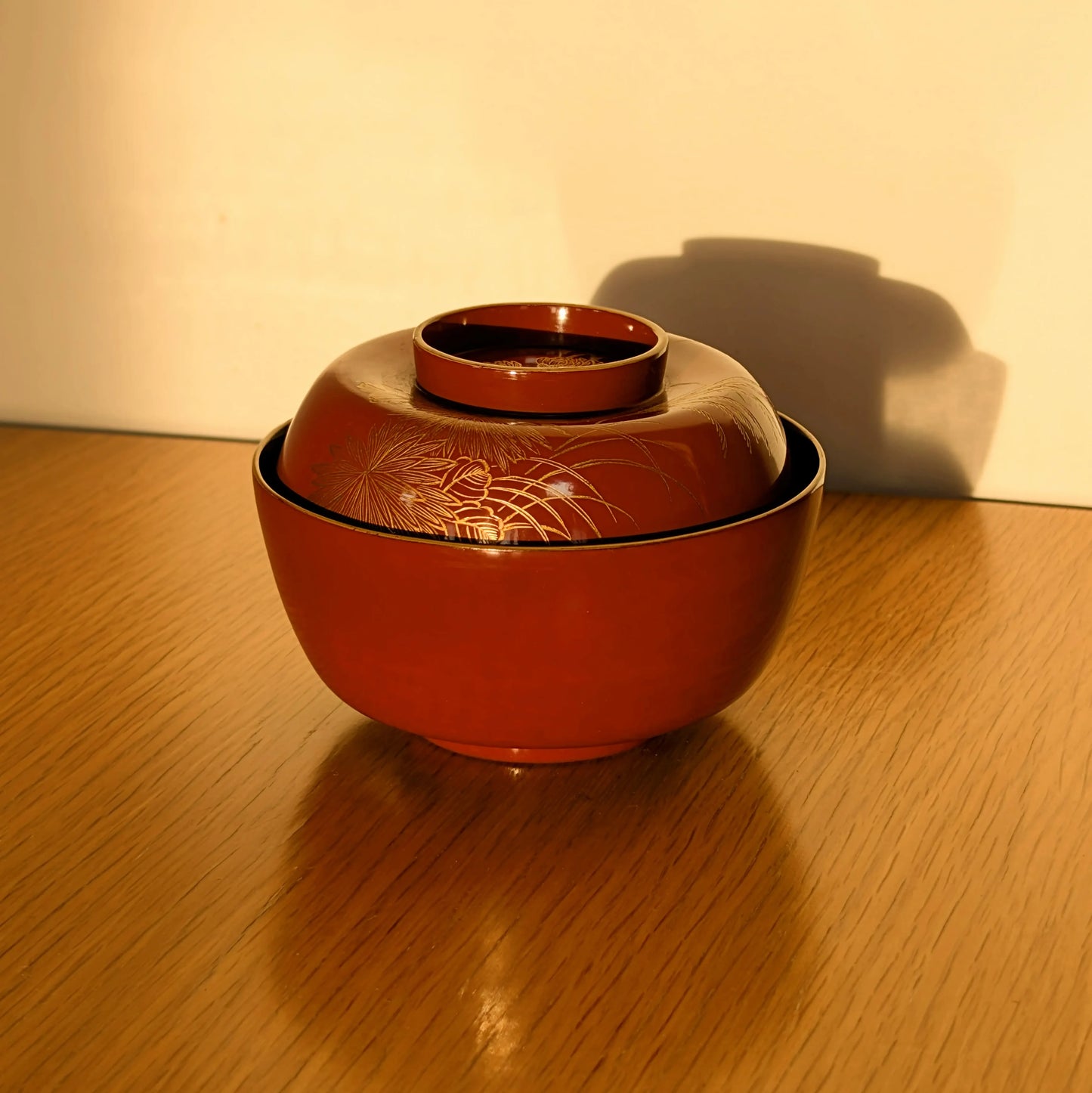 Bol japonais ancien en laque vermillon décoré en maki-e de chrysanthèmes et d’herbes susuki, photographié sous une lumière naturelle chaude.