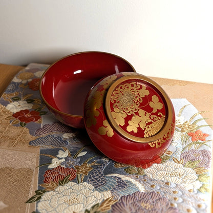 Bol japonais en laque urushi rouge et or présenté sur un textile japonais, avec couvercle orné de chrysanthèmes maki-e.