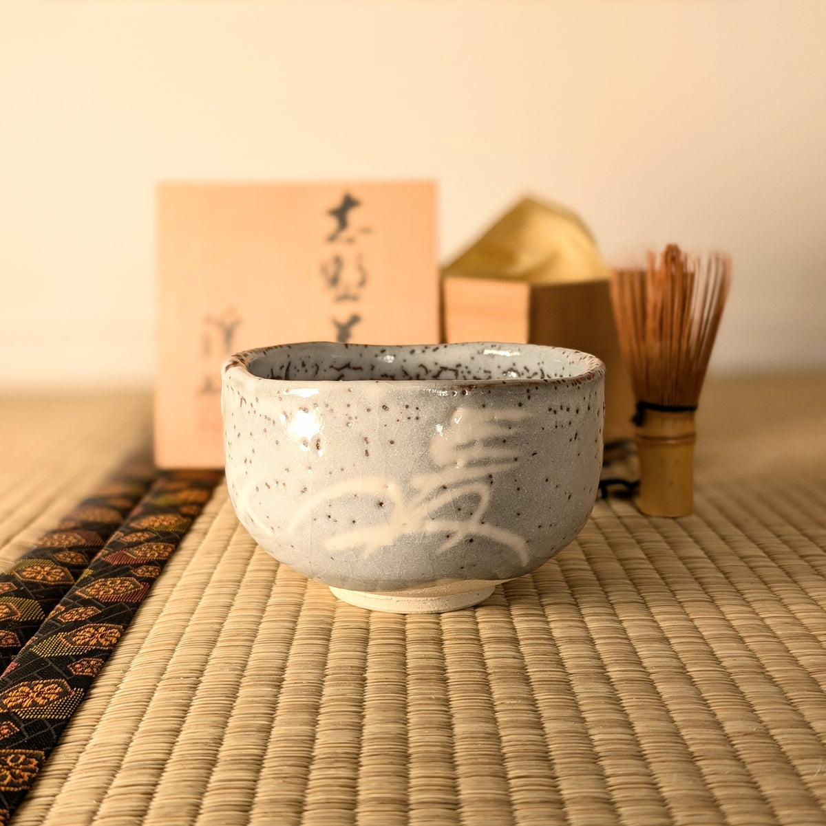 Bol à matcha japonais artisanal Nezumi-Shino mis en scène avec chasen et tomobako pour la cérémonie du thé chanoyu.