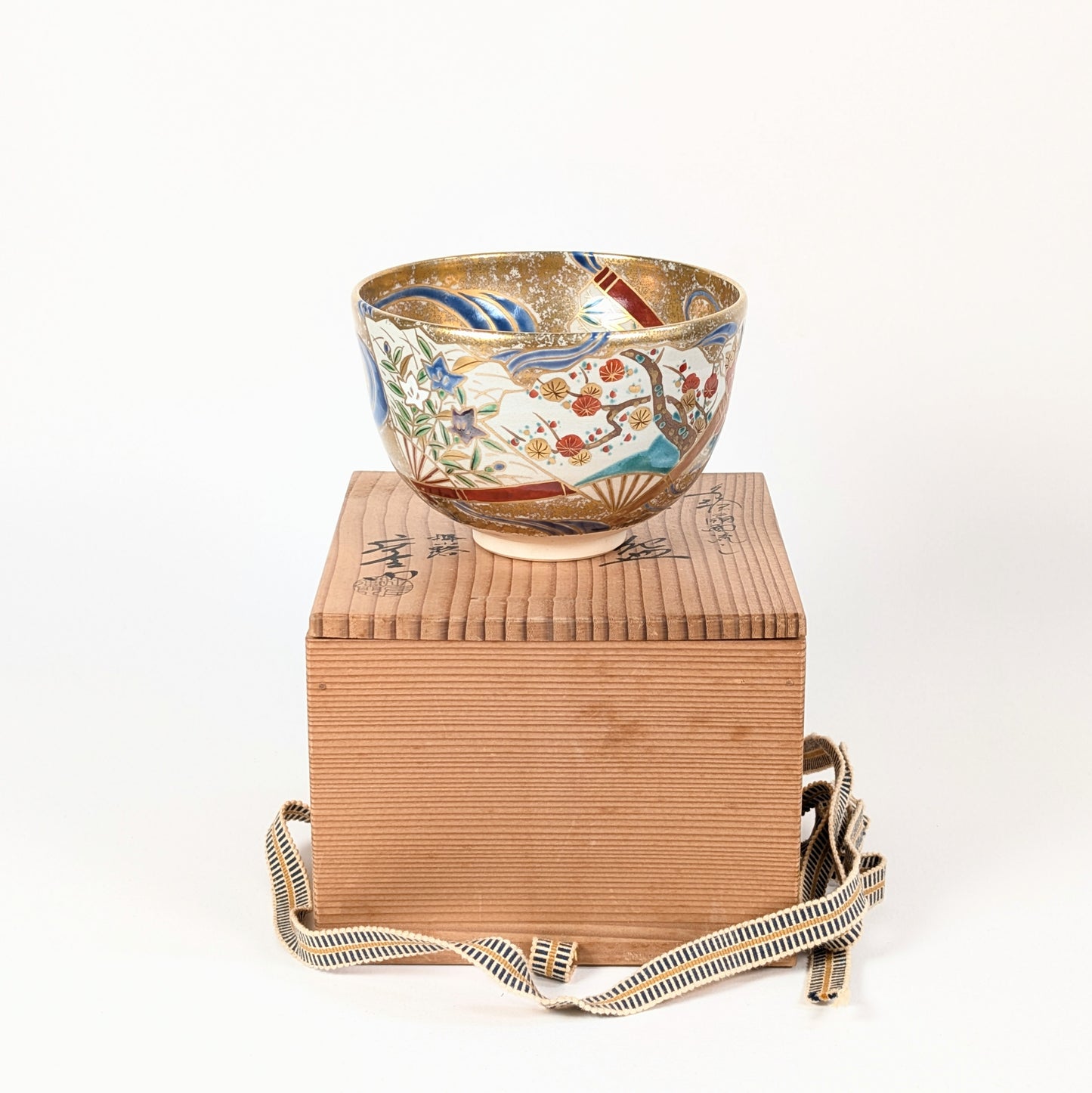 Bol à matcha japonais posé sur sa boîte en bois de kiri, avec décor d’éventails flottants, de prunier et de fleurs kikyō.