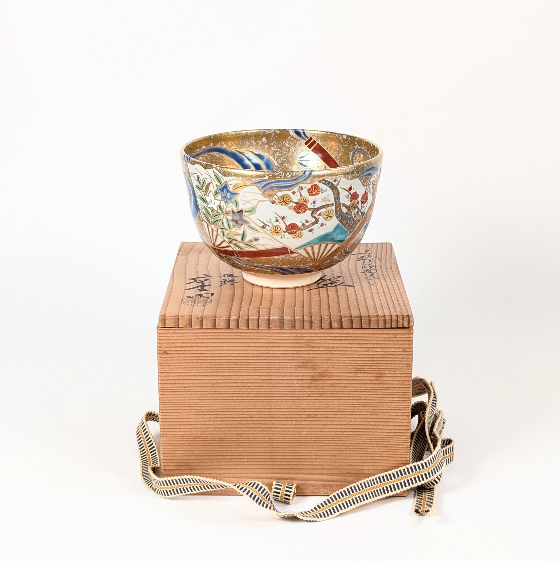 Bol à matcha japonais posé sur sa boîte en bois de kiri, avec décor d’éventails flottants, de prunier et de fleurs kikyō.