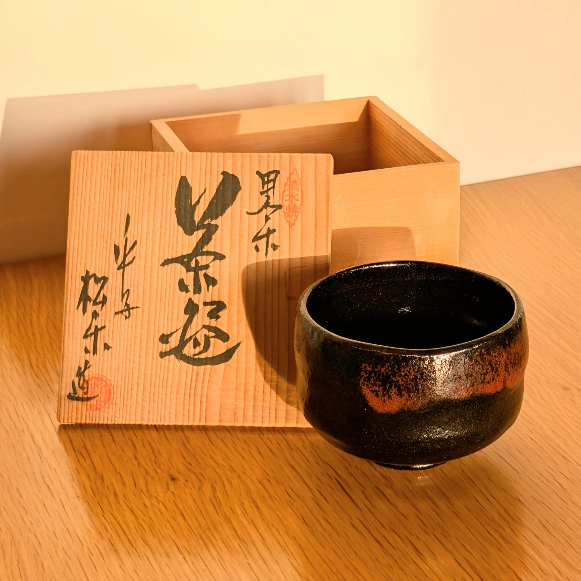 Chawan japonais en raku noir signé Sasaki Shōraku, avec tomobako calligraphiée, glaçure hi-iro et lumière chaude — artisanat de Kyoto.