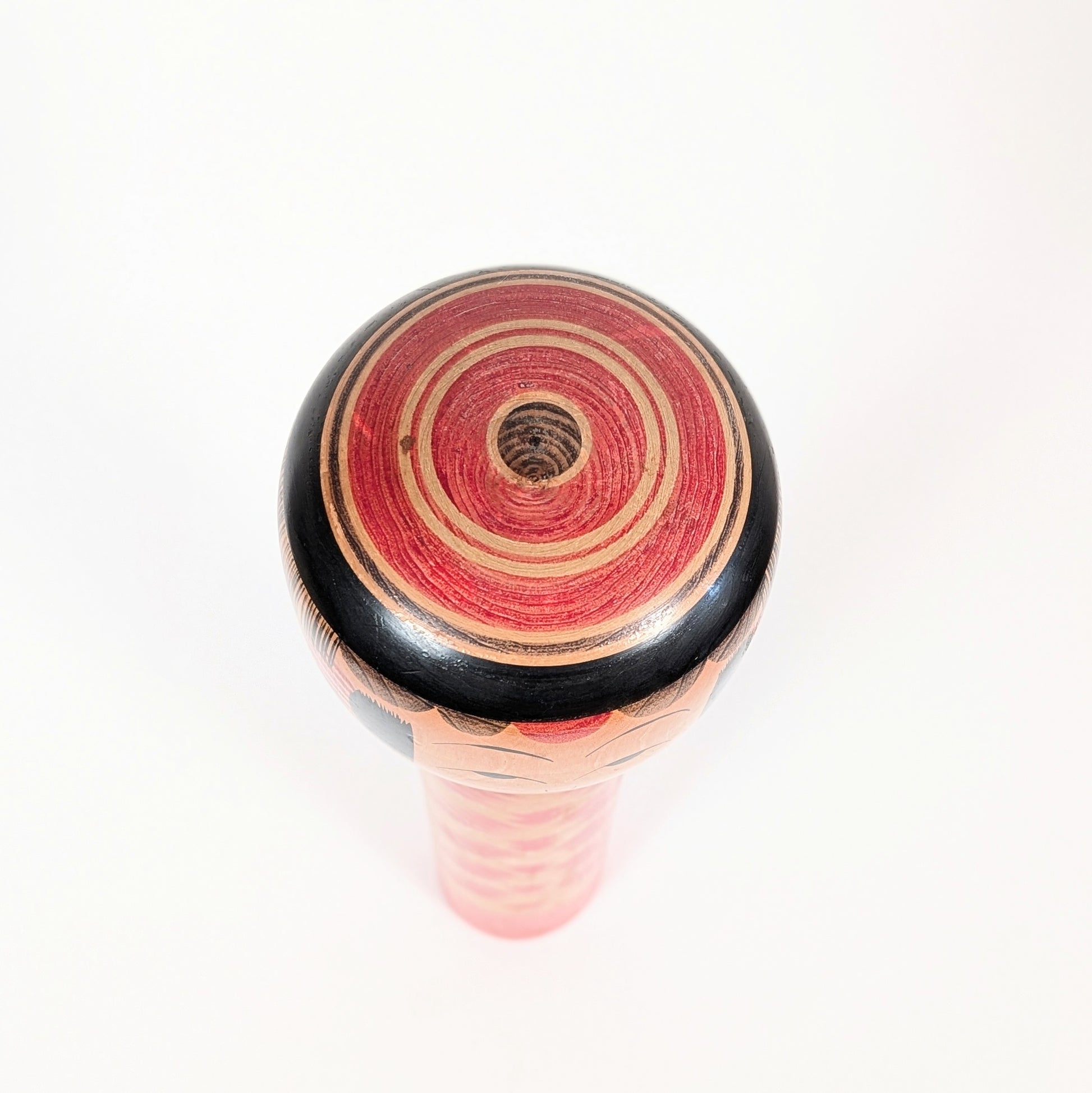 Vue du sommet de la kokeshi Yajirō avec coiffure janome, formée de cercles concentriques rouges, noirs et couleur bois.