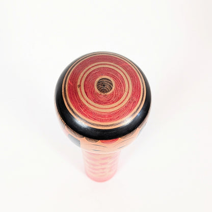 Vue du sommet de la kokeshi Yajirō avec coiffure janome, formée de cercles concentriques rouges, noirs et couleur bois.