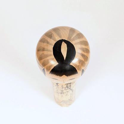 Vue du sommet de la tête de la kokeshi Shūhō, détail de la coiffure noire et du décor peint sur bois.