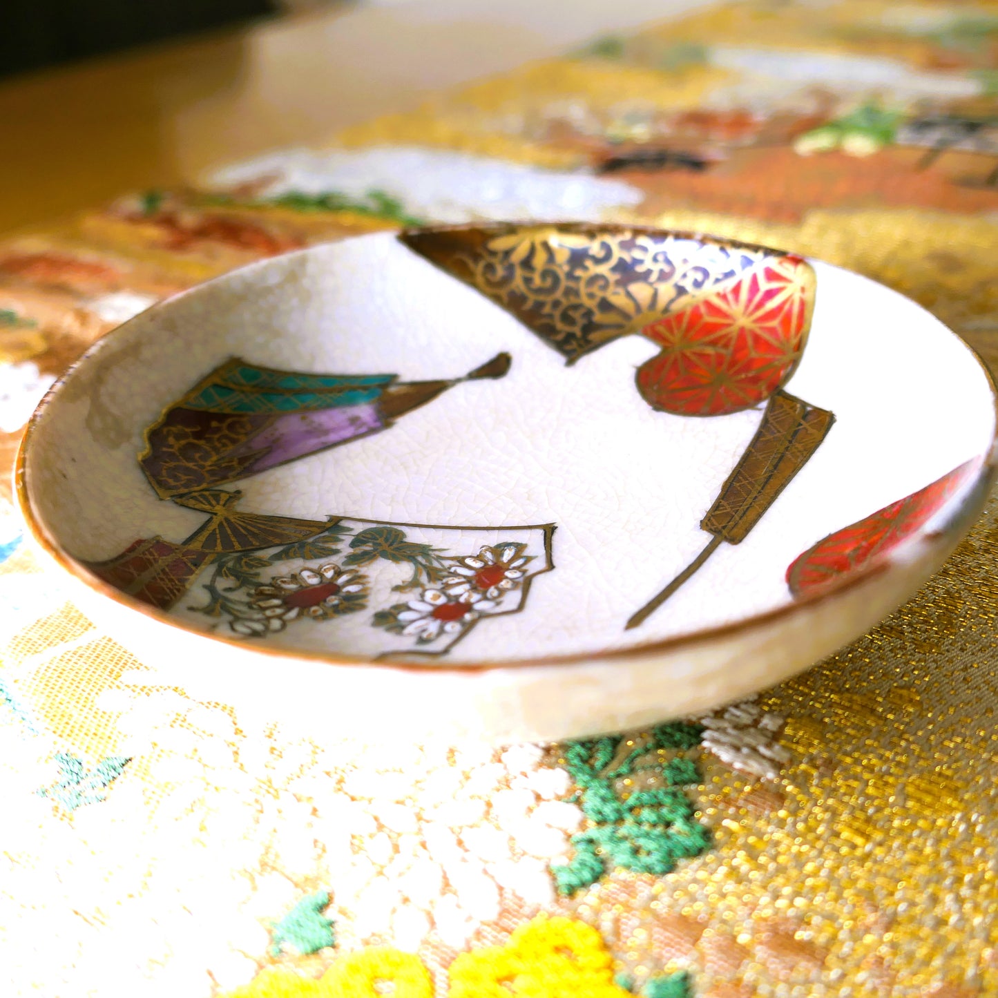 Coupelle japonaise en céramique Kutani ancienne, décorée de motifs d’éventails et posée sur un tissu d'un obi brodé traditionnel.