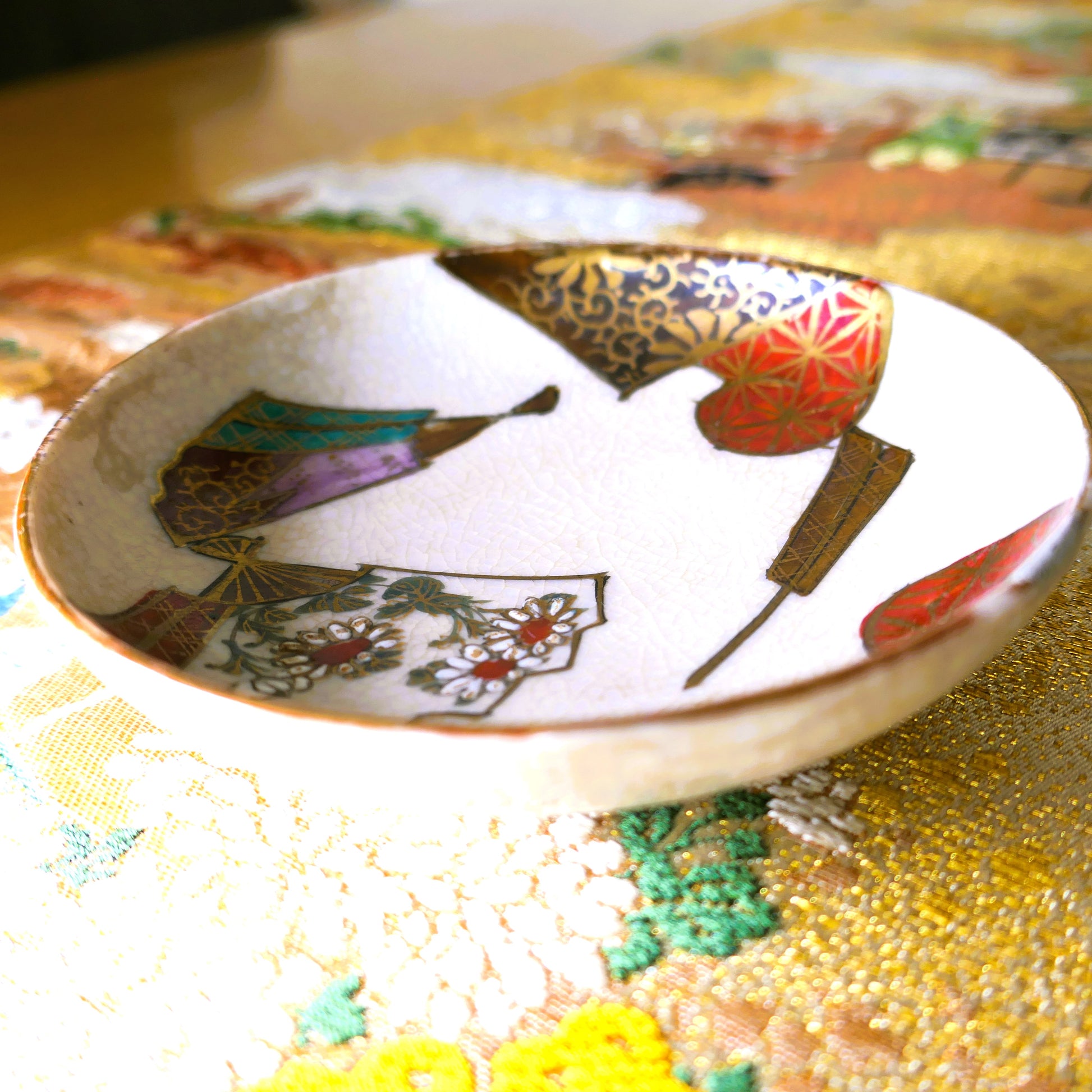 Coupelle japonaise en céramique Kutani ancienne, décorée de motifs d’éventails et posée sur un tissu d'un obi brodé traditionnel.
