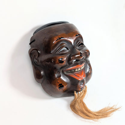 Vue de trois quarts d’un Daikoku-men japonais, laque rouge-brun sur fond noir et barbe en fibres végétales.