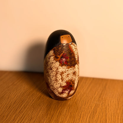 Kokeshi japonaise vintage Shôzan présentée sur une surface en bois, dans une lumière chaude mettant en valeur ses érables d’automne.