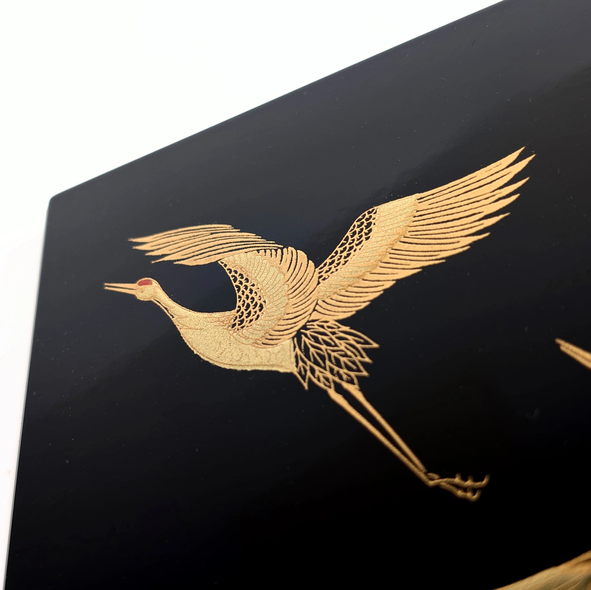 Gros plan sur une grue tsuru dorée en maki-e, ailes déployées sur fond de laque noire.