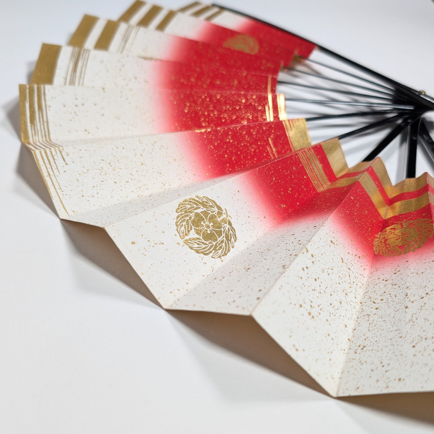 Objet d’art décoratif japonais : éventail en papier cartonné rouge, blanc et or, orné de kamon, pris en vue plongeante.