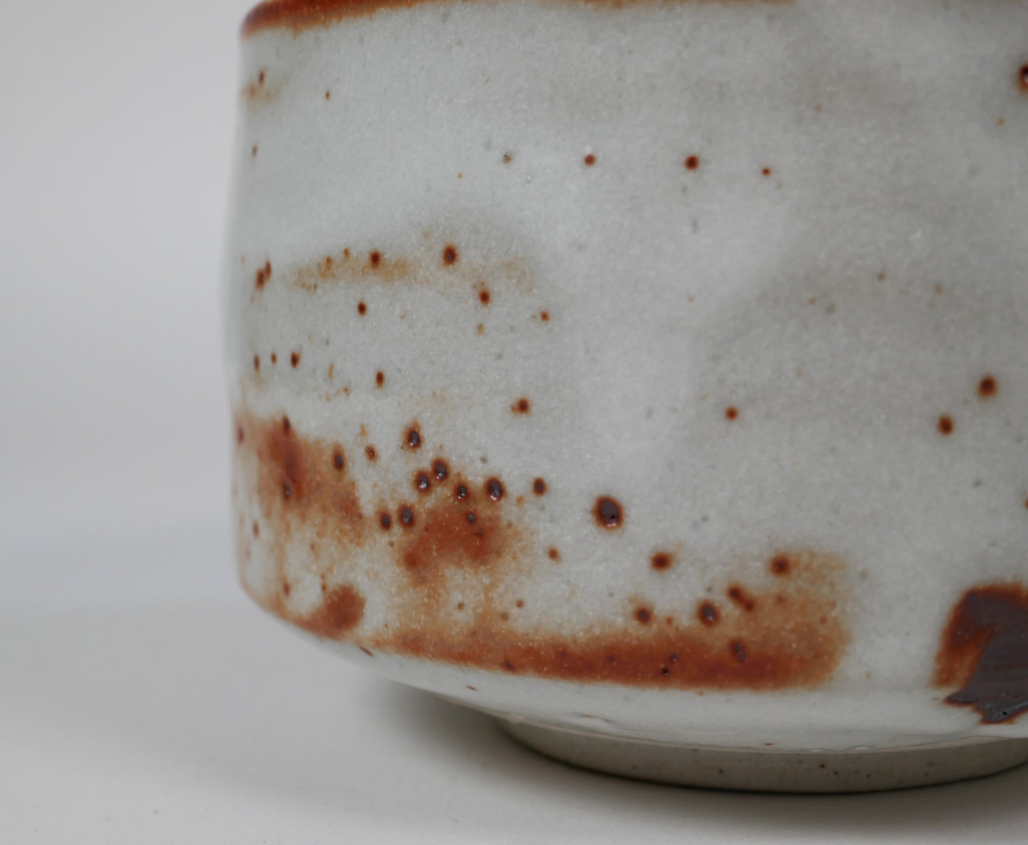 Gros plan sur l’émail Beni Shino avec points ferrugineux, glaçure blanche texturée et marques de cuisson d’un bol à thé japonais artisanal.