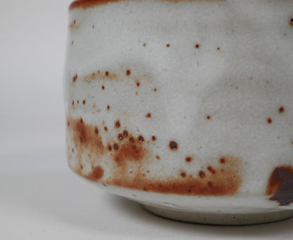 Gros plan sur l’émail Beni Shino avec points ferrugineux, glaçure blanche texturée et marques de cuisson d’un bol à thé japonais artisanal.