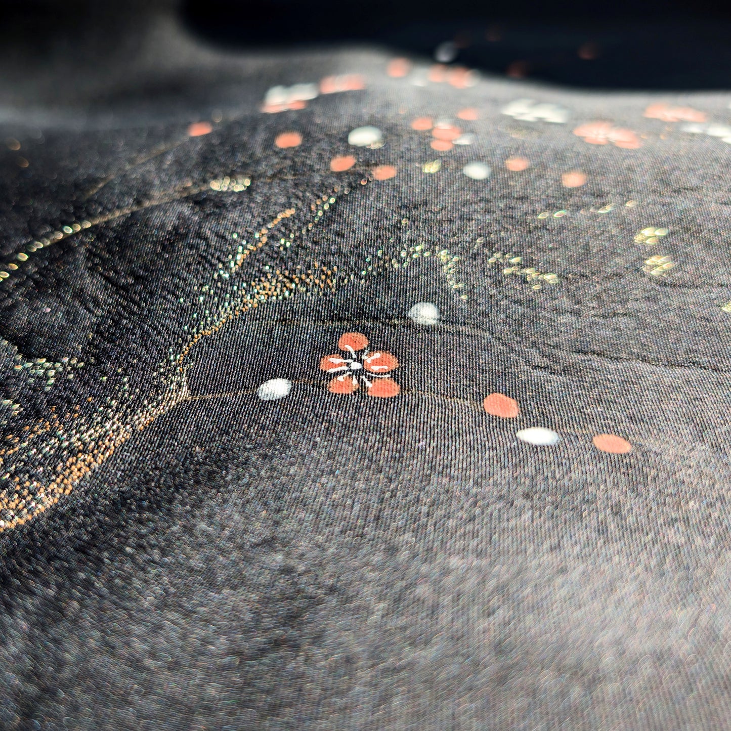 Macro de la soie noire discrètement brillante d’un haori japonais, avec texture rinzu en relief, fleurs ume rouges et blanches et branches dorées.